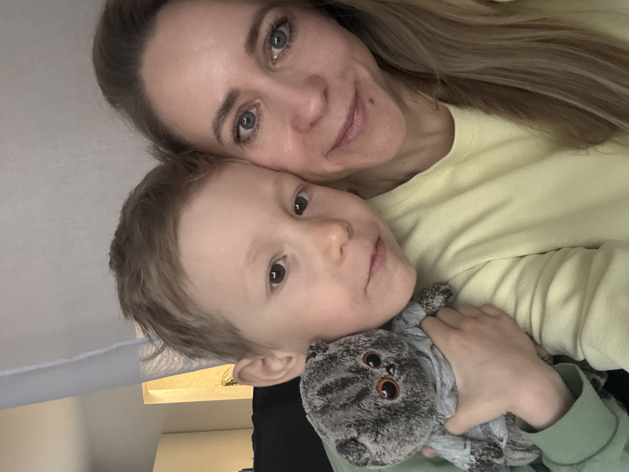 A selfie-style photograph shows a woman and a young child. The woman has fair skin, blue eyes, and long blonde hair. She is wearing a light yellow shirt. The child has fair skin, light brown hair, and is holding a gray plush cat toy. The child is wearing a green long sleeve shirt and a black shirt can be seen under the child’s. Based on the information given, I am unable to verify the location is in Chaikovsky, Russia.