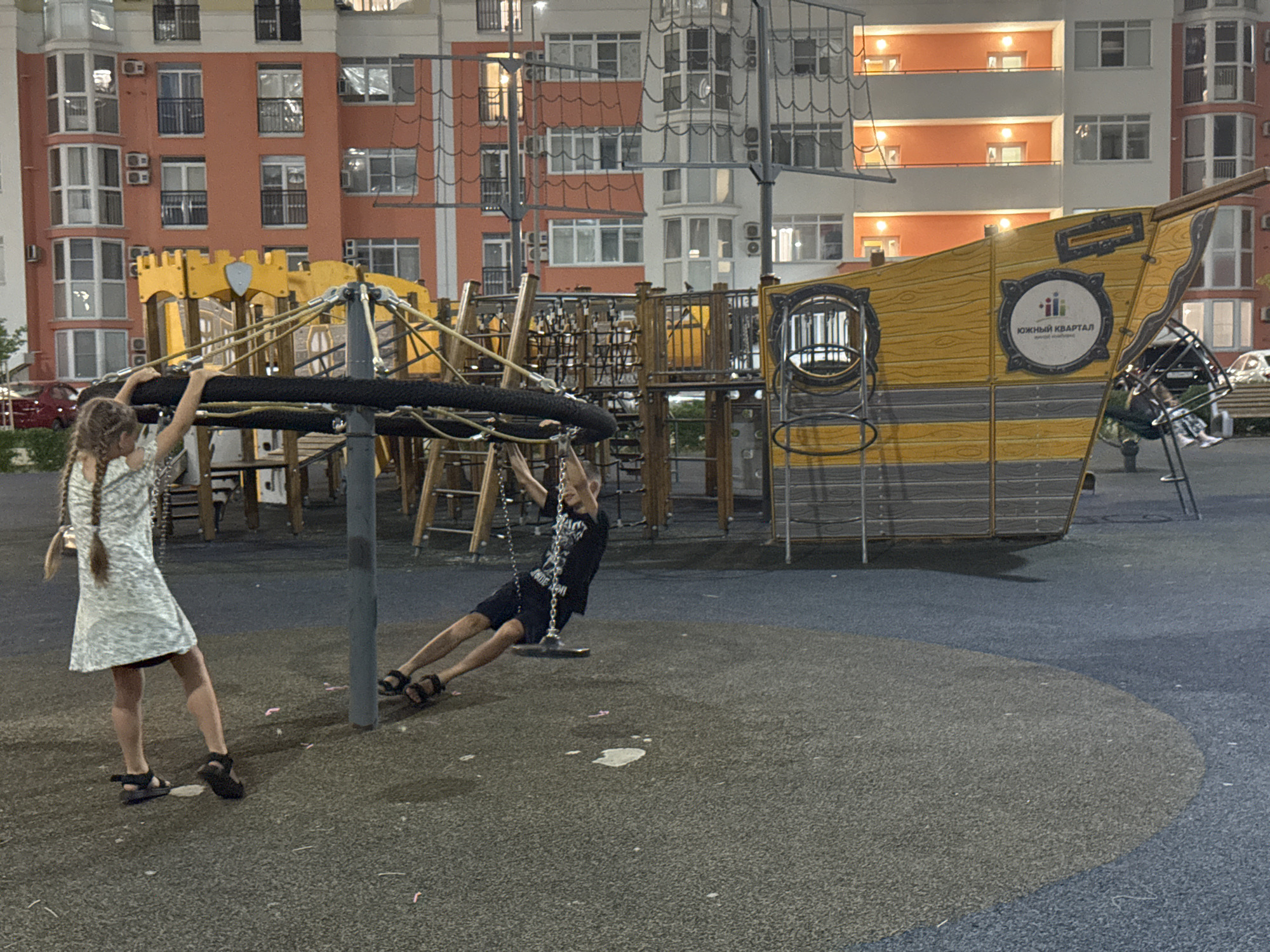 The image depicts a playground with children on it in the city of Anapa, Russia. In the foreground, a young girl with braided hair wearing a light-colored dress and dark sandals stands on a black rubberized surface, her left hand gripping a dark bar. To her right and further back, a boy in a black t-shirt and dark shorts is swinging on a chain swing. He is positioned on the same rubberized surface, his legs extended. In the background, a large, yellow, ship-themed play structure dominates the scene, with a logo on its side that appears to read "Южный Квартал." Behind the playground, multi-story residential buildings with balconies and windows are visible, illuminated by interior lights, suggesting it is dusk or evening. The overall environment appears to be a modern, urban playground.