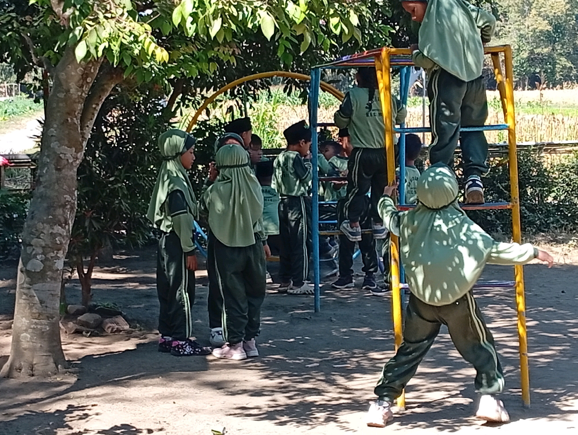 In Miritpetikusan, Indonesia, a group of children in matching green and dark green uniforms are gathered around a colorful playground structure. A large tree with textured bark stands to the left, casting shadows on the sandy ground. The playground features yellow and blue metal bars, with a red circular arch. Several children are climbing on the structure, including one child standing on top of the yellow ladder and another on a lower rung. Other children are observing from the ground. The environment appears to be outdoors, with foliage and sunlight suggesting a daytime scene.