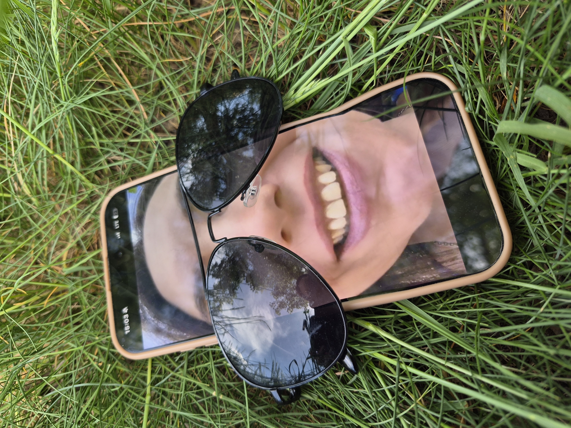 The image shows a cell phone with a photo of a smiling face on its screen, placed on a bed of green grass. A pair of black aviator sunglasses rests on top of the phone. The reflection in the sunglasses suggests an outdoor environment with trees and sky. The phone displays a time of 15:03. The location is Unknown.