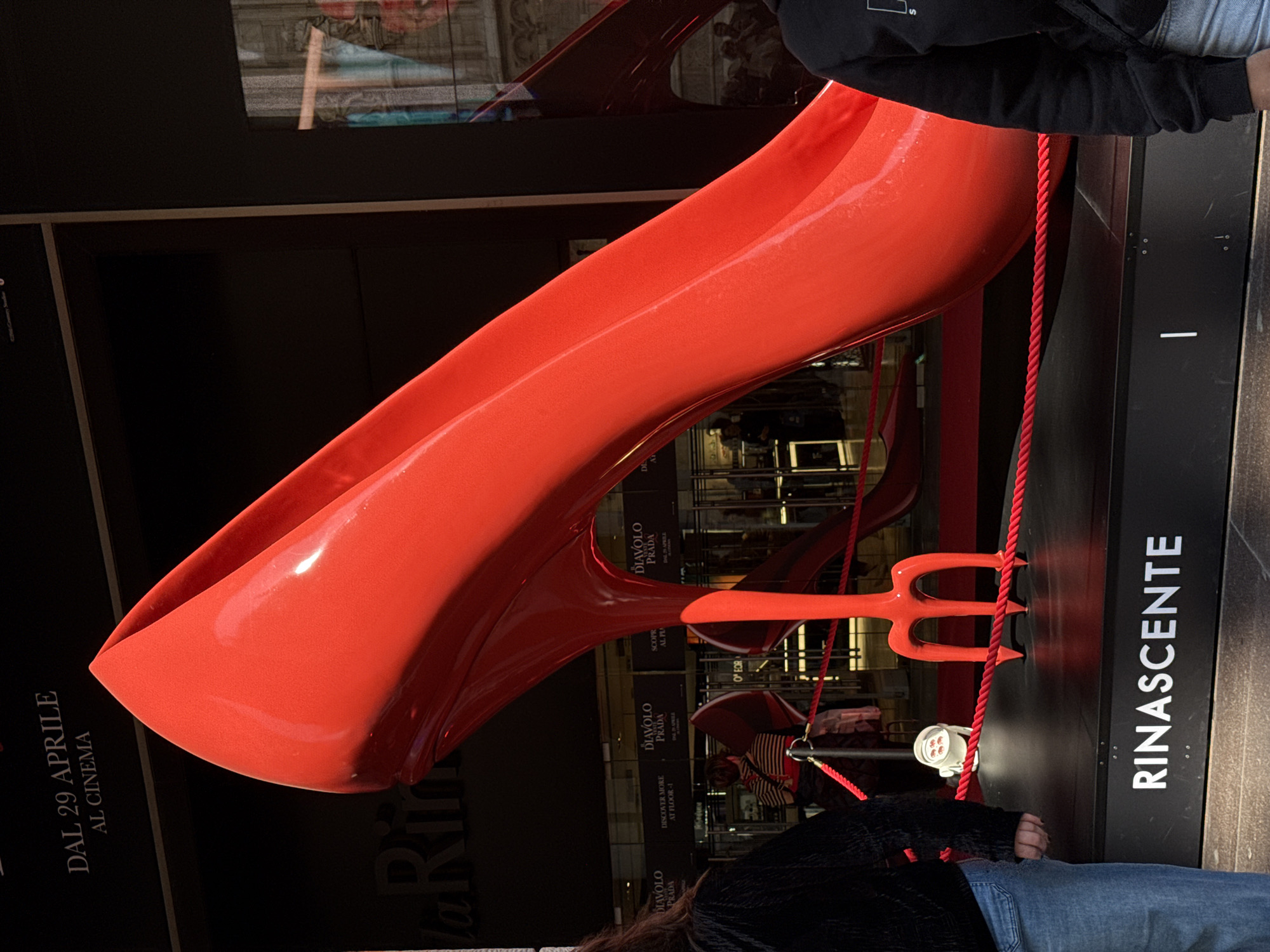 Here is a detailed description of the image:
The image captures an outdoor advertisement for the movie "Diavolo Prada," which translates to "Devil Wears Prada" or "The Devil Prada" – an Italian adaptation of the original name. The advertisement is affixed to the side of the "Rinascente" department store, located in Milan, Italy.
The advertisement is quite striking and features a large, oversized, shiny red high-heeled shoe sculpture. The shoe appears to be positioned dynamically, as though it's in mid-stride or taking a step. Beside the shoe is a similarly large red trident. The "Rinascente" branding is clearly visible, indicating the location of the advertisement. The date "DAL 29 APRILE AL CINEMA" suggests that the movie was or will be playing in cinemas from April 29th onwards.
People are seen in the image, though they are partially cut off, suggesting they were simply passing by. They appear to be wearing casual clothing and are likely pedestrians.
The location, being outdoors and in front of a department store in Milan, indicates a commercial environment, likely a busy urban area. The image has been taken at a medium angle, capturing both the details of the advertisement and the nearby surroundings.