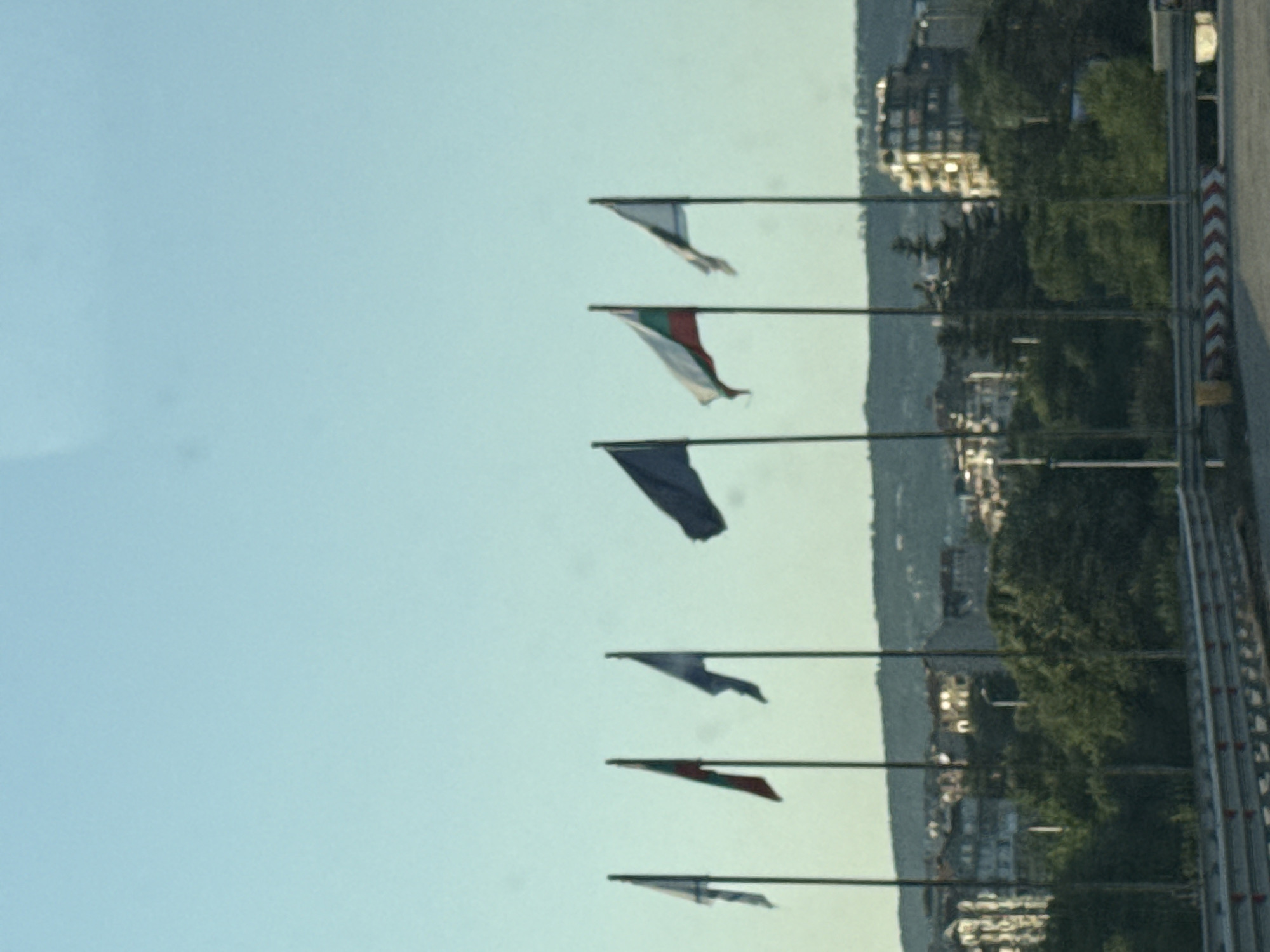 This media file features a series of flags flying against a pale blue sky. The flags are of various colors and designs, with some appearing to be solid colors and others featuring patterns or emblems. They are mounted on poles, suggesting an outdoor setting. In the background, there is a blurred landscape that includes buildings nestled amongst lush green trees. A road or pathway with a red and white striped barrier is visible on the right side of the frame. The image has a somewhat hazy or washed-out quality, and it is difficult to discern specific details of the flags or the landscape. There is no visible text in the image. The lighting suggests it might be daytime, possibly late afternoon due to the softness of the light. The overall impression is one of a somewhat serene, open-air scene, possibly a park, a race track, or a scenic overlook.