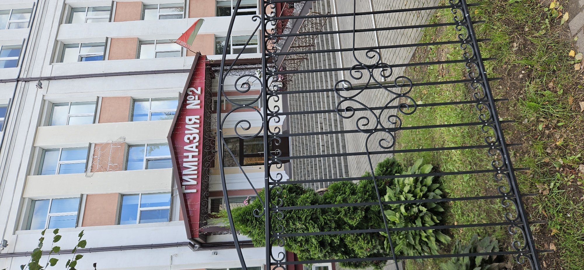 This is a medium shot, eye-level, outdoor photograph of the entrance to Gymnasium No. 2 in Vitsebsk, Belarus. The building is a multi-story structure with a facade of cream and light orange panels, and white trim around numerous windows. The entrance features a red, gabled awning with the text "ГИМНАЗИЯ №2" in white letters. Decorative strings of lights are draped along the underside of the awning. A Belarusian flag, red and green, flies from a pole to the right of the entrance. In front of the building, a black wrought-iron fence runs horizontally across the lower portion of the frame. To the left of the entrance, behind the fence, is a neatly trimmed evergreen tree next to a patch of leafy green plants. Beyond the entrance, a wide set of concrete stairs leads up to a dark-colored doorway. The ground in front of the stairs is paved with light-colored interlocking bricks. The overall impression is that of a well-maintained educational institution on a clear day.