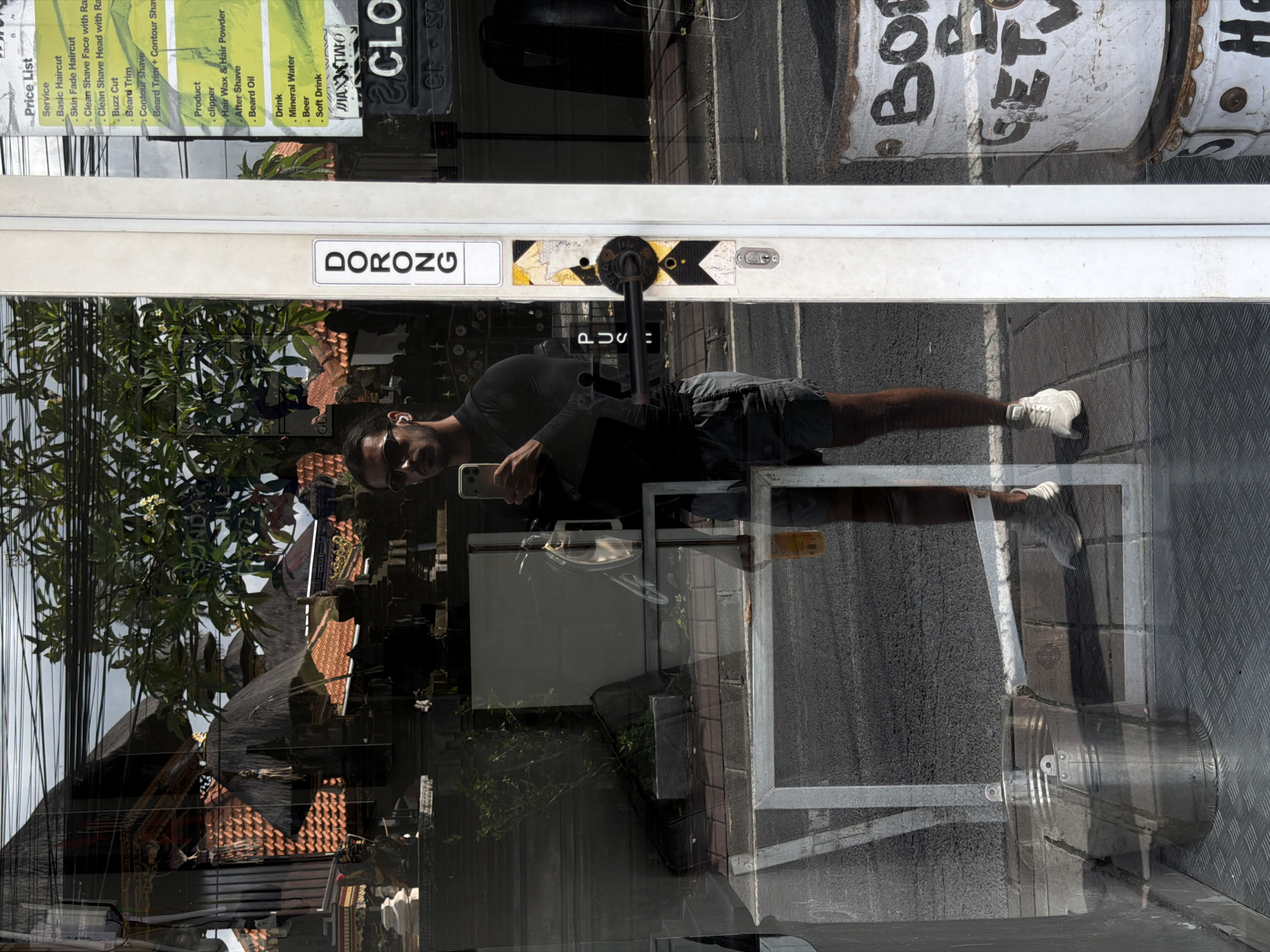 A man stands in front of a glass door and window with a price list on the left edge in North Kuta, Indonesia, holding a cell phone up in his right hand as if taking a photo. He has sunglasses on his face and is facing toward the viewer. He has short dark hair and a dark t-shirt. He is wearing grey shorts and light-colored sneakers.
The glass door has "DORONG" written in white letters on a black background above a door handle. A "PUSH" sign is directly below the handle.
Reflections in the glass show a tiled roof, tree branches, and electrical lines.
To the right are several metal cans with painted graffiti designs.