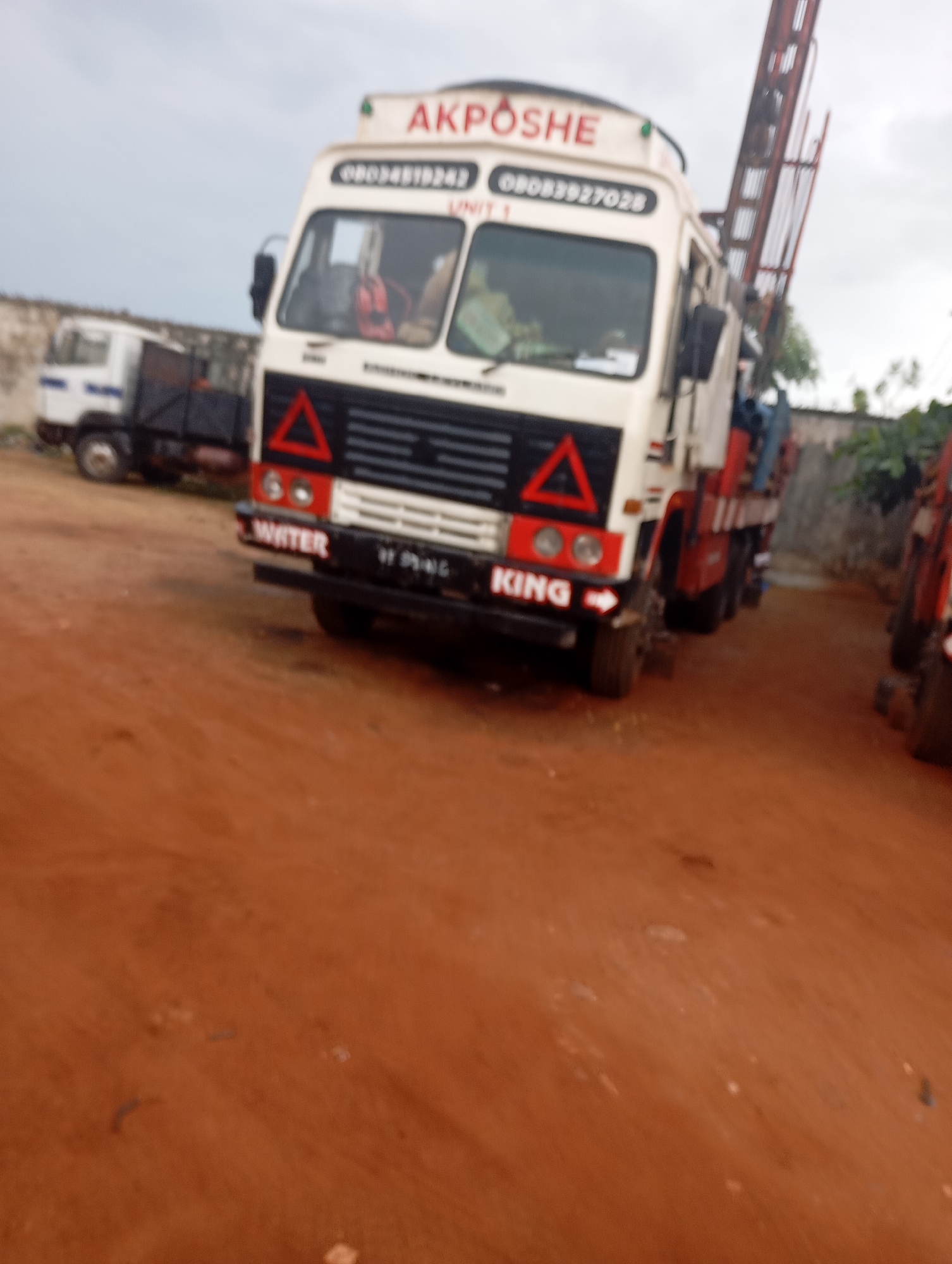 The image depicts a drill rig truck with "AKPOSHE" written in red lettering on its front. Below this, the phone number "08034519243" and "08033927028" are visible, along with "UNIT 1". The truck has a white and red exterior and a black grill. In front of the truck, the words "WATER" and "KING" are printed in white letters on a black surface, with an arrow pointing to the right. There is another truck partially visible in the background to the left. The ground appears to be made of reddish-brown earth, consistent with the arid soil common in Shendam, Nigeria. The sky is overcast. The scene suggests an industrial or service setting in Shendam, Nigeria, likely related to water drilling or supply operations.