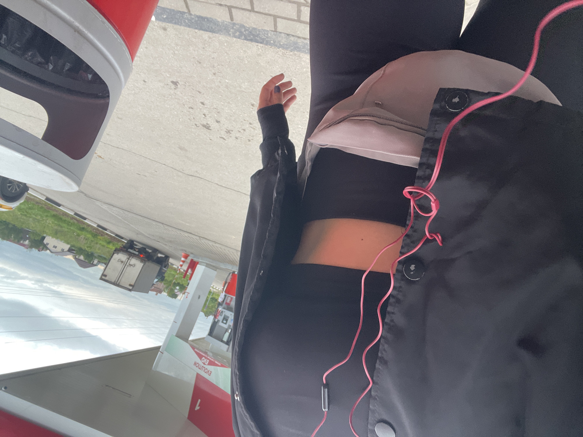 The image shows a person at a gas station, likely in Molokovo, Russia, based on the surrounding environment. The person is wearing black clothing and a light-colored hip bag. Pink headphones are visible. In the background, there is a gas station canopy, gas pumps marked with "EVOLUTION EVO" and the number "1", and a truck driving on a road. Houses are seen in the distance under a cloudy sky. A red and gray trash bin is also visible.