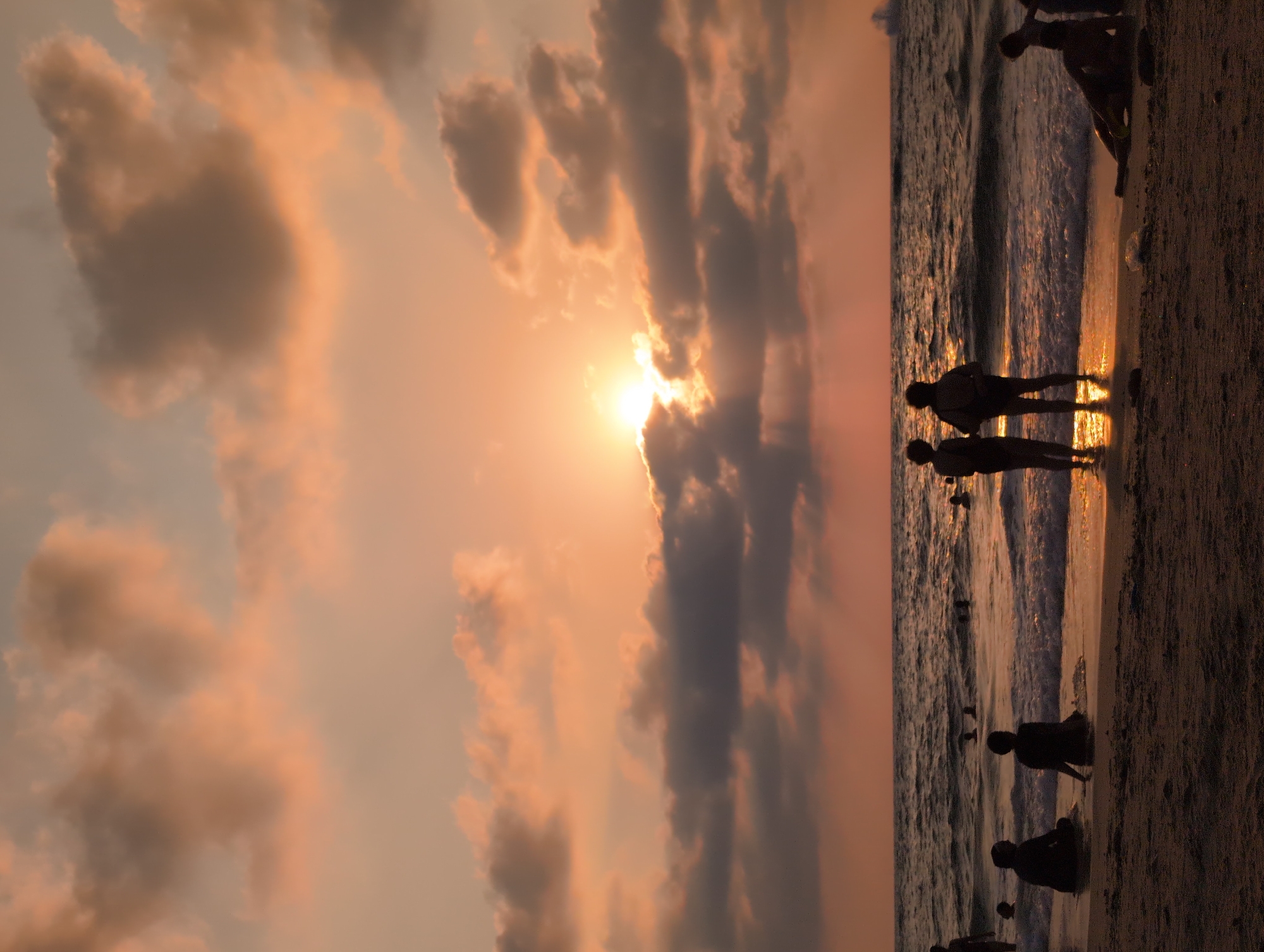 The image depicts a serene sunset scene at Palmachim Beach in Israel. The sky is painted with warm hues of orange and pink, with scattered clouds partially obscuring the setting sun, casting dramatic sunbeams onto the water. The sea is calm with gentle waves lapping at the shore. Silhouetted figures of people are scattered across the beach and in the shallow water, enjoying the last moments of daylight. Some are standing, others are sitting, and a few are wading in the sea. The foreground is a sandy beach with some discarded plastic items, suggesting recent human activity. In the distance, a faint outline of a ship can be seen on the horizon, adding to the vastness of the seascape. The overall atmosphere is peaceful and tranquil.
