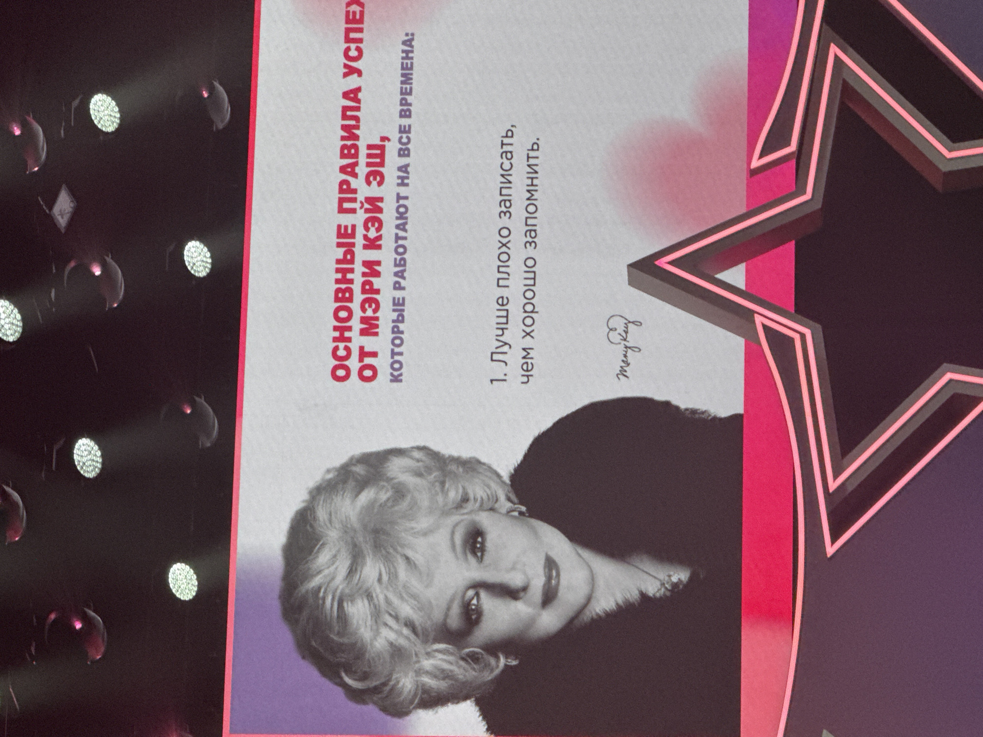 The image shows a poster with a black and white portrait of Mary Kay Ash, founder of the Mary Kay Cosmetics company. She is looking directly at the camera with her signature bouffant blonde hair and makeup. The text on the poster in Russian reads "BASIC RULES OF SUCCESS FROM MARY KAY ASH, WHICH WORK FOR ALL TIME:" and then "1. It is better to write down poorly than to remember well." Below the text, there is a signature that appears to say "Many Key". The poster is framed by a pink and purple border with a stylized star shape at the bottom. The background of the poster is a light gray, with some pinkish-red accents that appear to be a gradient from the border. The overall presentation suggests a motivational or educational message, likely for a sales or business audience. There is no information in the image to indicate its location in Moscow, Russia.