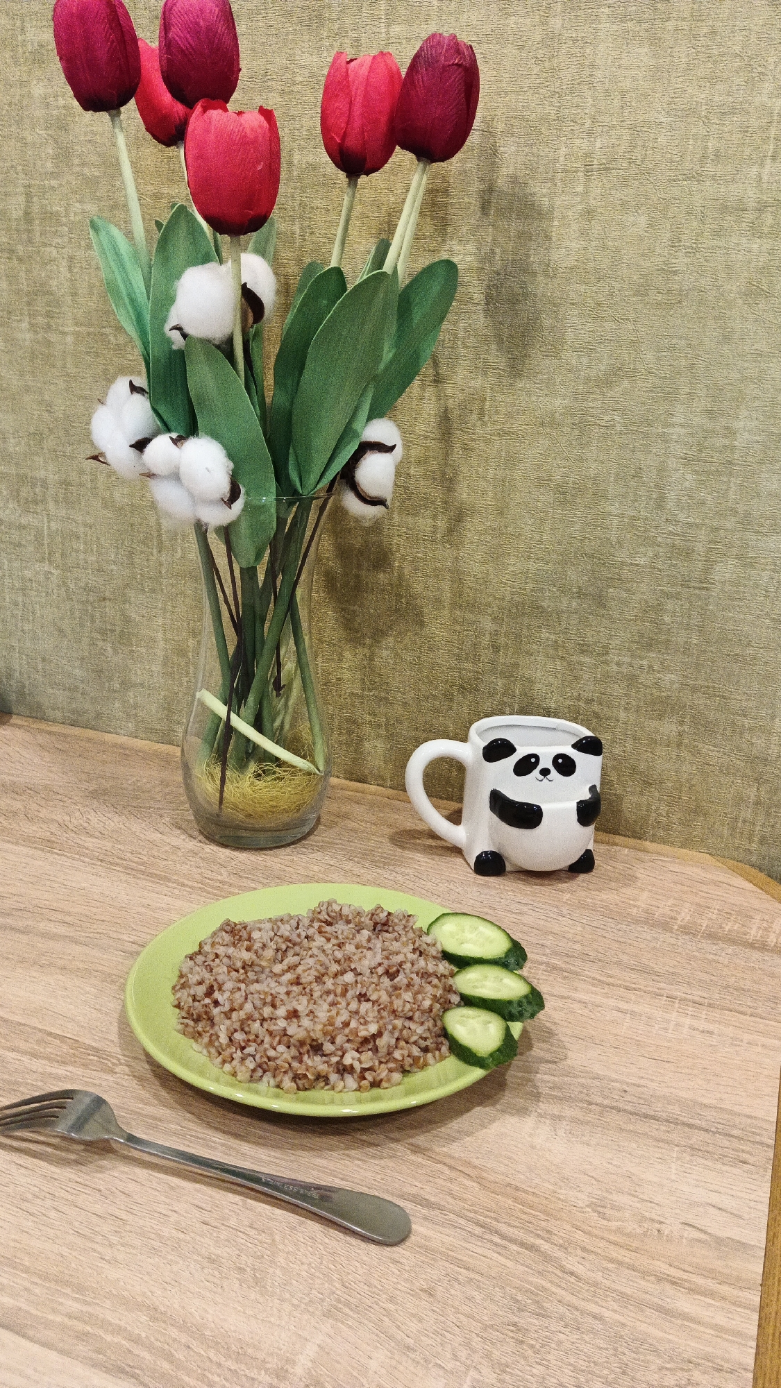 In Kryvyi Rih, Ukraine, a still life arrangement is set on a wooden table. A clear glass vase contains artificial red tulips and white cotton bolls, with green stems and leaves. To the right of the vase, a white ceramic mug shaped like a panda is placed. In the foreground, a light green plate holds a portion of cooked buckwheat with three slices of cucumber arranged alongside. A silver fork rests in front of the plate. The background features a textured wall in a muted green hue.
