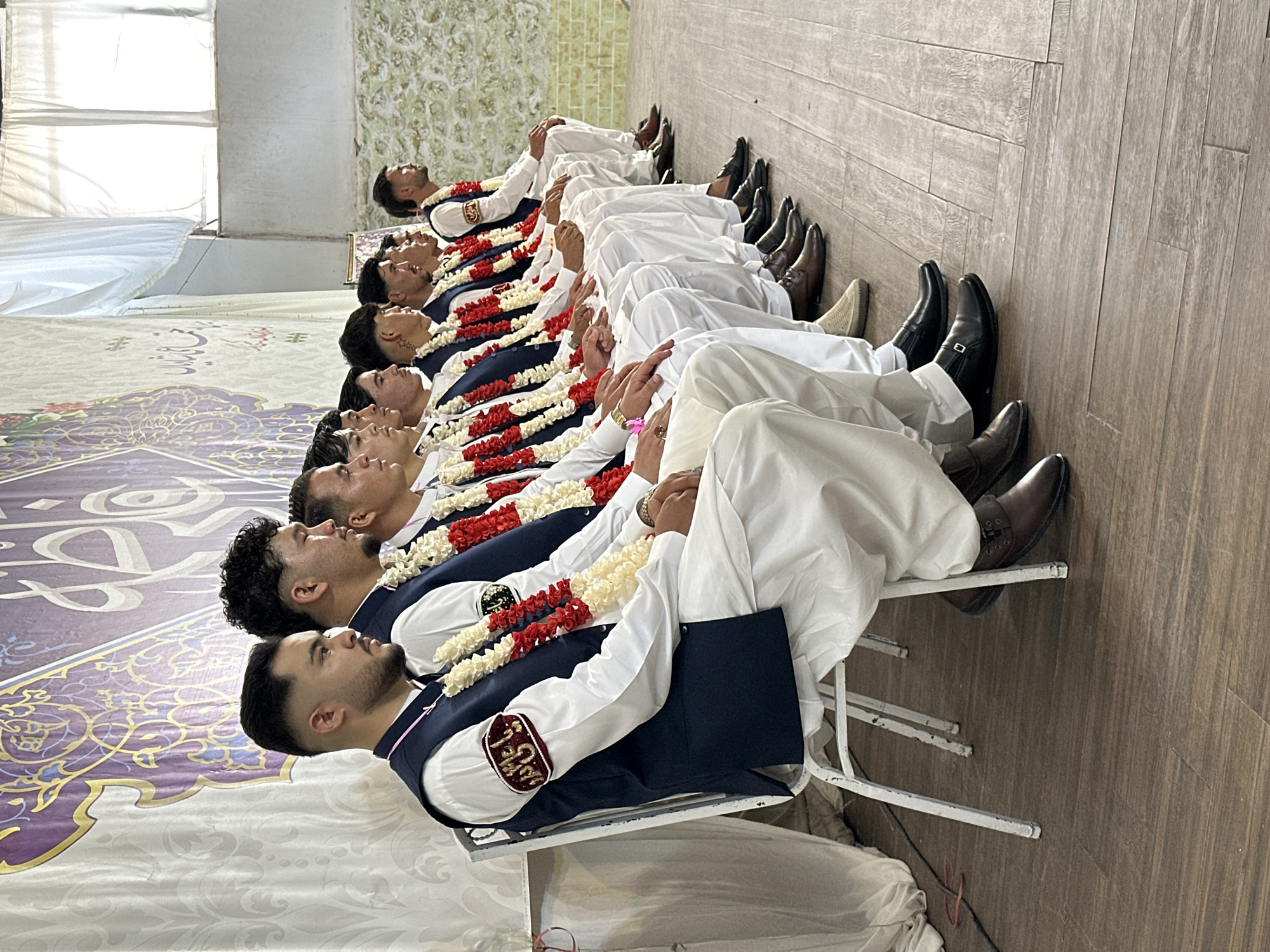 A photograph captures a row of men seated on chairs in Quetta City Tehsil, Pakistan. They wear matching white outfits with navy blue vests and garlands of red and white flowers. Most have dark hair and are looking up. Their shoes range from brown leather to black dress shoes. The room has wood flooring, and a carpet with an ornate purple and gold design is visible on the left side of the frame. White fabric is draped on the sides of the room.