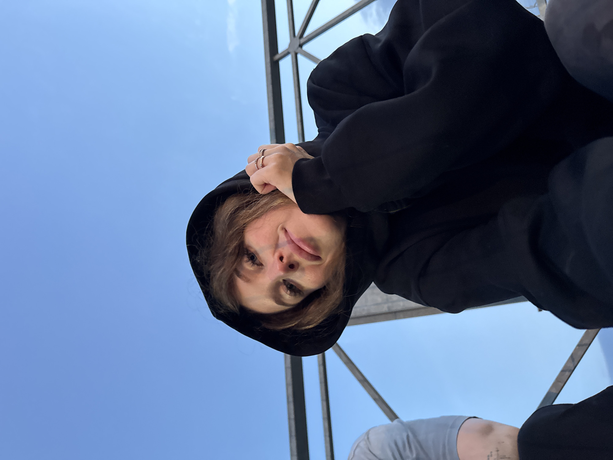 The media file shows a low-angle, upside-down close-up of a person wearing a black hoodie with the hood up. The person's face is partially visible, with their tongue sticking out slightly. They are wearing several rings on their fingers, which are resting near their head. The background is a clear blue sky with a glimpse of a modern architectural structure, possibly a bridge or walkway, made of dark metal. The lighting suggests it is daytime, likely afternoon given the brightness of the sky. The overall mood appears playful and candid. The scene is likely outdoors, possibly in an urban or park-like setting given the architecture. There is no visible text in the image. The person's pose and expression suggest a sense of fun or silliness. Another person is partially visible at the bottom right of the frame, wearing a grey t-shirt with a tattoo on their arm.