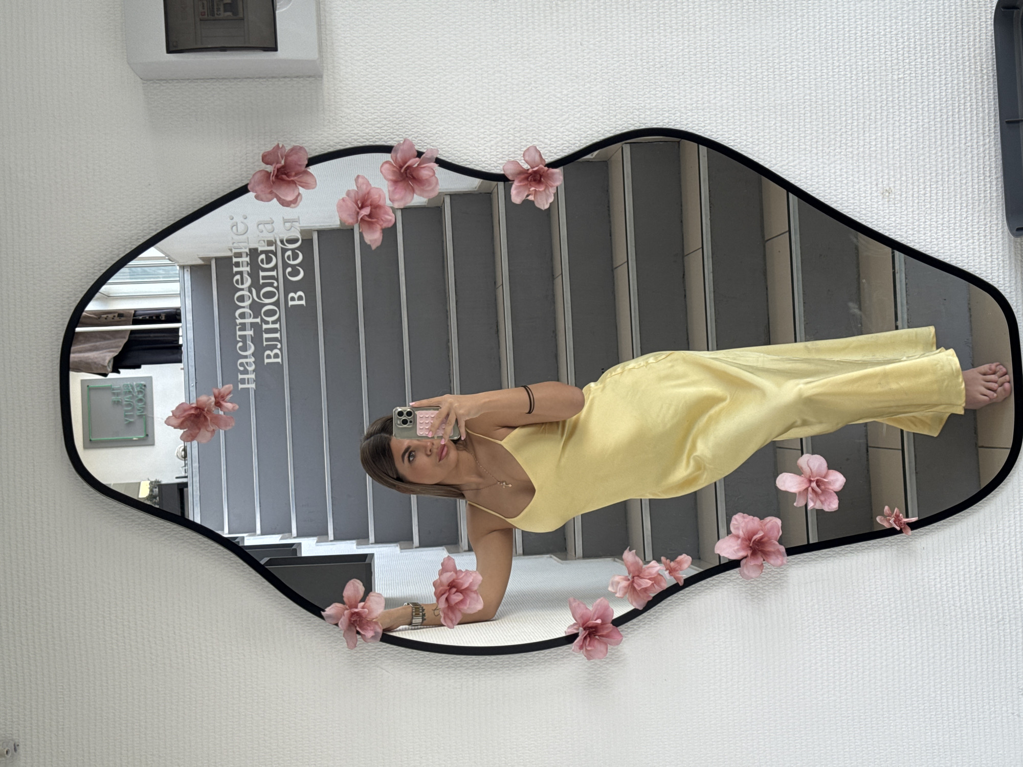 A young woman in a yellow dress is reflected in a decorative mirror adorned with pink flowers. The mirror is mounted on a white wall. The reflection shows a staircase behind her. In the mirror, text in Cyrillic is visible. The woman is holding a smartphone and taking a selfie. The location is the city of Chaikovsky, Russia.