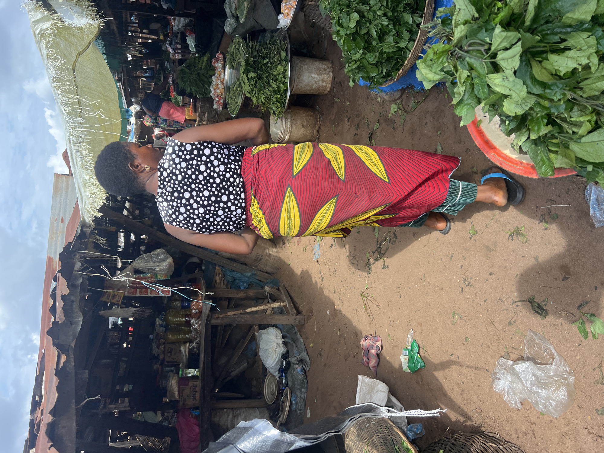An outdoor market scene shows a woman standing in front of a stall in Unknown, Unknown. The woman, with dark hair, is wearing a black and white polka dot top and a long red skirt with yellow leaf-like patterns. She is wearing sandals on her feet. To her right, there are various vegetables and leafy greens displayed in baskets and buckets. The ground is dirt, with scattered debris and plastic bags. To her left, a weathered stall is visible, stocked with various items, including bottles, packages, and other goods. The stall has a makeshift roof of metal sheeting and fabric. The sky is visible in the background.