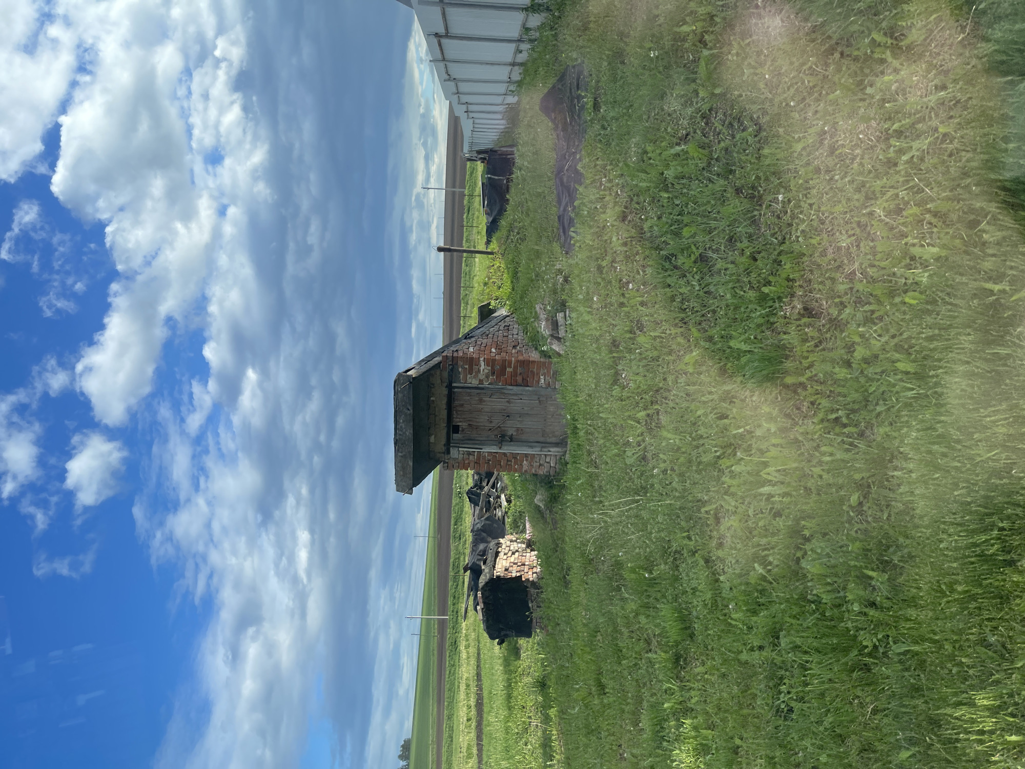 The image depicts an outdoor scene in a grassy area, likely in the outskirts of a city, possibly in Shemetovo, Russia, given the prompt's mention. A small brick and wood structure, resembling an outhouse or shed, stands prominently in the foreground. Debris, possibly collapsed bricks or other construction materials, are scattered around the base of the structure. The foreground is dominated by green grass and weeds, suggesting an unmaintained or rural setting. In the background, there is a field or cultivated area and the sky is visible with a mix of blue and white clouds. A fence or wall structure runs along the edge of the field. There are no people visible in the image. The overall impression is one of neglect and abandonment.