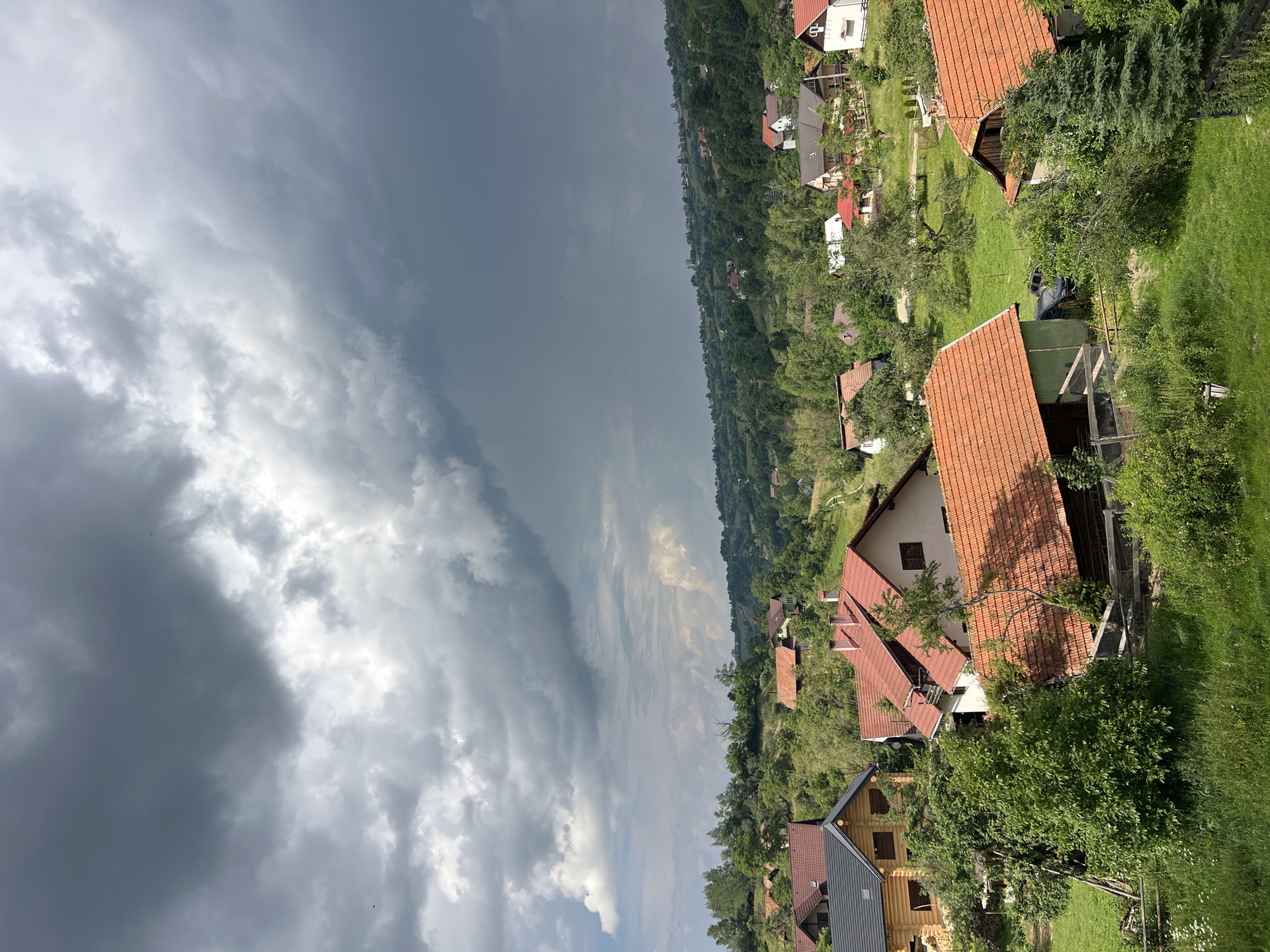 A high-angle, eye-level shot shows a cluster of houses with red-tiled roofs nestled among rolling green hills under a dramatic, cloudy sky. The houses vary in size and style, with some featuring wooden exteriors and others rendered in lighter colors. Several trees and dense foliage surround the buildings, and a winding dirt road is visible in the distance. The sky is filled with dark, heavy clouds, suggesting an impending storm, contrasted by patches of lighter sky and distant, lighter clouds. The overall scene depicts a rural or semi-rural residential area in a hilly landscape.