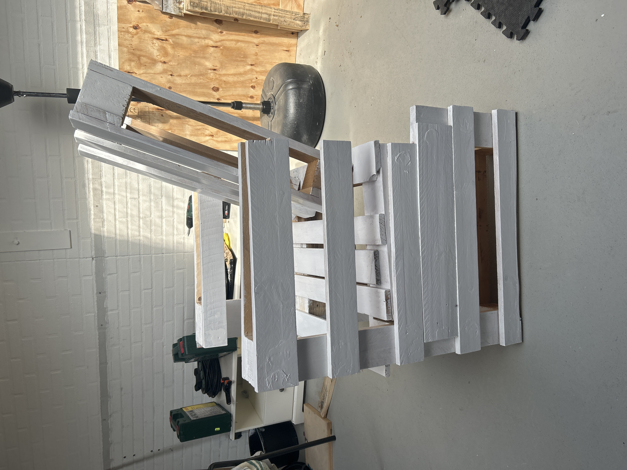 In Lisse, Netherlands, a white painted pallet chair is positioned in what appears to be a workshop or garage. The chair is made from disassembled and repurposed wooden pallets, with the backrest angled upwards. The chair is partially painted white, with some areas of raw wood visible, suggesting it is either a work in progress or a rustic design. The floor is a concrete surface. In the background, a white brick wall is visible, along with various items including two green toolboxes, coiled cables, a white cabinet, a large black punching bag, and sheets of plywood. Sunlight streams in from an unseen window, casting shadows across the scene.