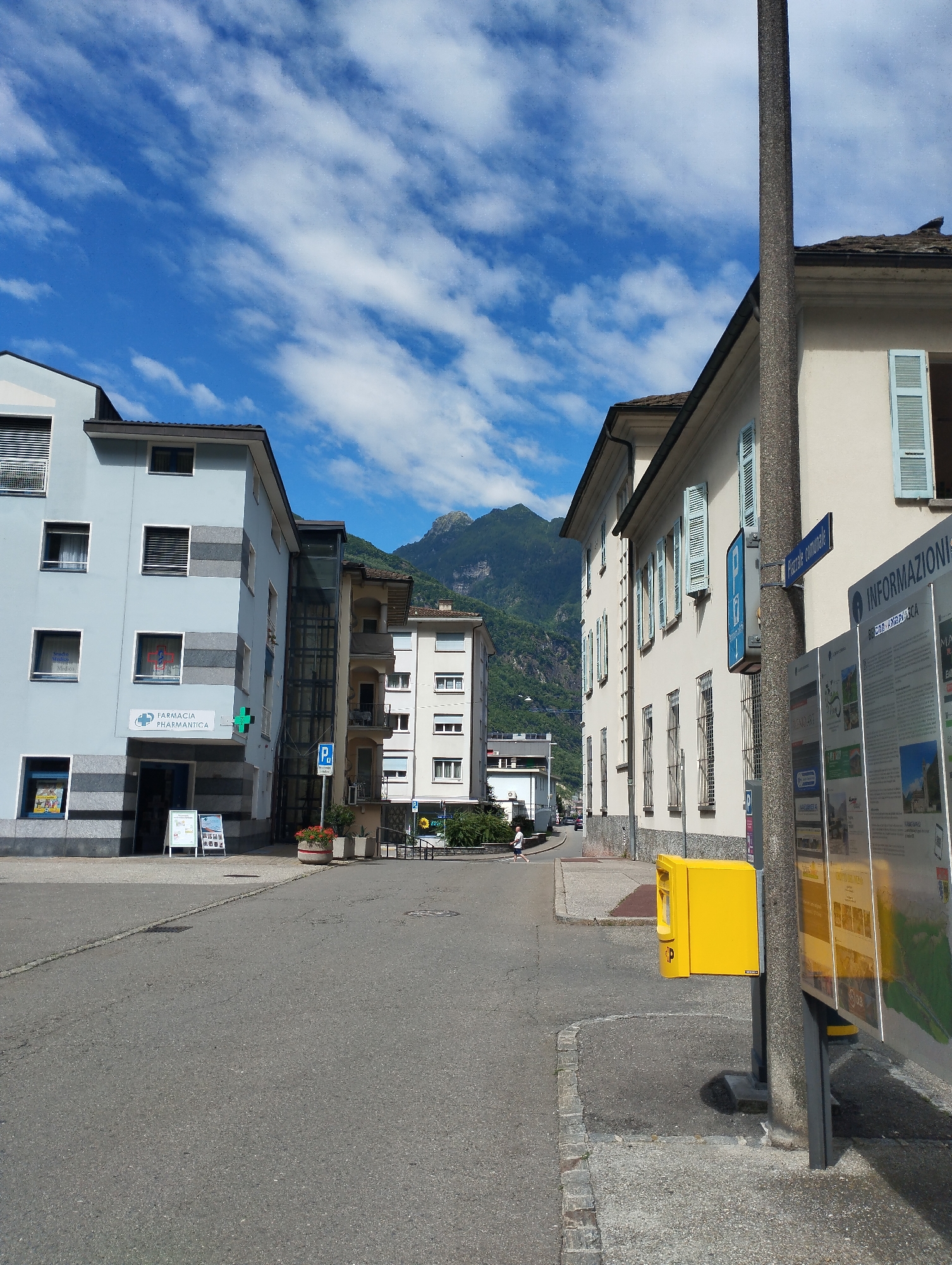 A street scene in a town with mountains in the background, under a bright blue sky with scattered white clouds. On the left, a light blue building features a pharmacy with a green cross logo. In the foreground, the asphalt street is empty except for a few distant pedestrians. To the right, a cream-colored building has many windows with light blue shutters. A lamppost stands prominently in the right foreground, next to a bright yellow public utility box and a large informational kiosk displaying maps and text.