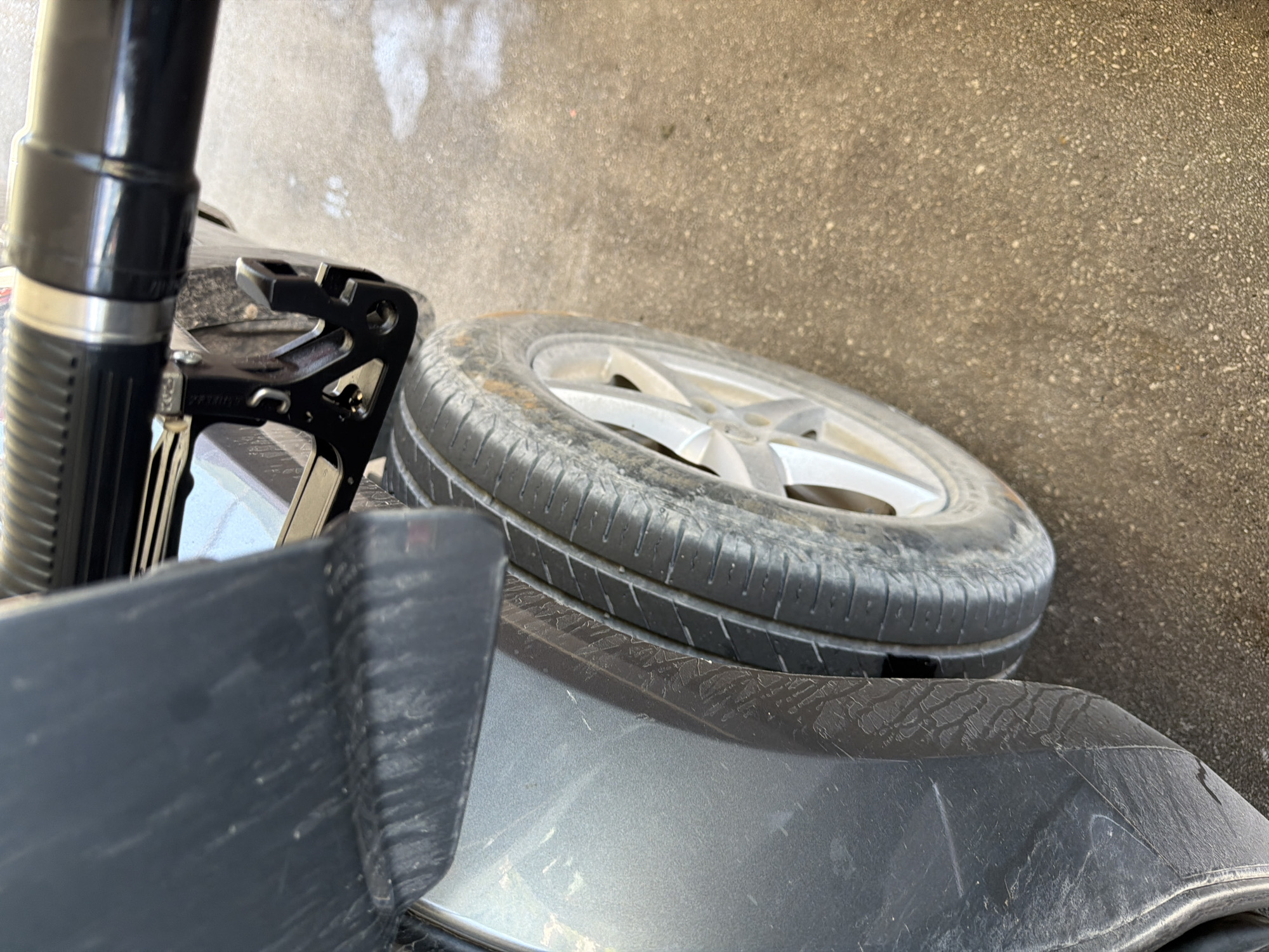 In Meljine, Montenegro, a close-up shot focuses on the side of a dark-colored vehicle, specifically its rear tire and part of the fender. A black fuel nozzle is inserted into the fuel door of the car. The tire is moderately worn and shows some dirt. The surrounding ground appears to be asphalt or concrete, with a light scattering of what might be sand or dirt. The lighting suggests an outdoor, daytime scene.