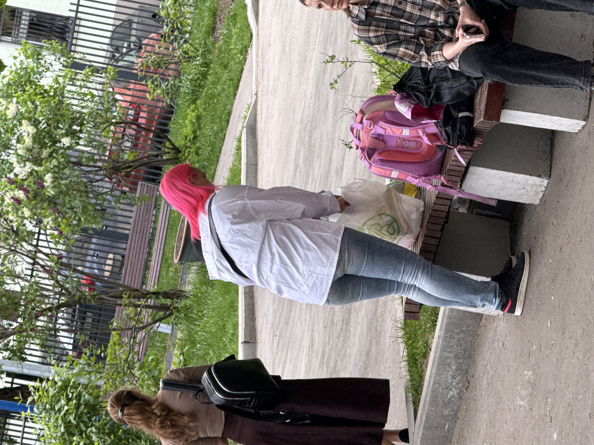 The image shows a street scene, likely in a residential area of Moscow, Russia. Three people are visible. One person with bright pink hair is standing and wearing a white shirt, light blue jeans, and black shoes. A person seated on a bench is wearing a plaid shirt and dark pants, looking at a mobile phone. Another person wearing a brown dress with a black and white backpack over their shoulder is at the bottom left. A pink backpack is on the bench next to the seated person. The ground is paved, with greenery along the edges of the walkway. A bench is in the background, along with a red car. A tall metal fence and buildings are visible beyond the street.