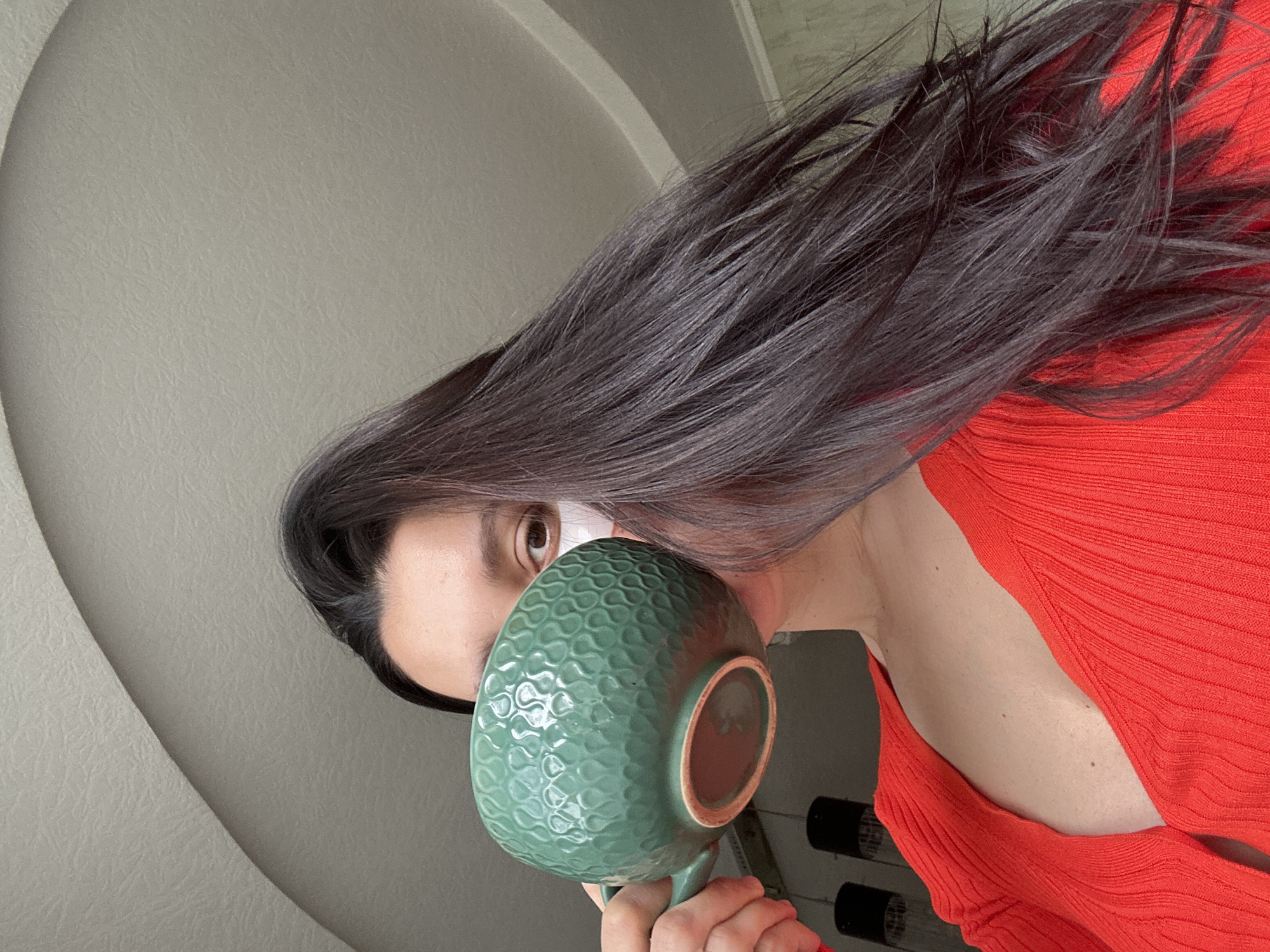 A woman with long, dark hair wearing a red top is holding a large green mug with a textured surface. The mug obscures most of her face, with only her eye and forehead visible. She appears to be indoors, with a neutral-colored wall and ceiling in the background. Below her shoulder, and in the background, is a black object.
It is not possible to determine the location as Chaikovsky, Russia based on the provided image.