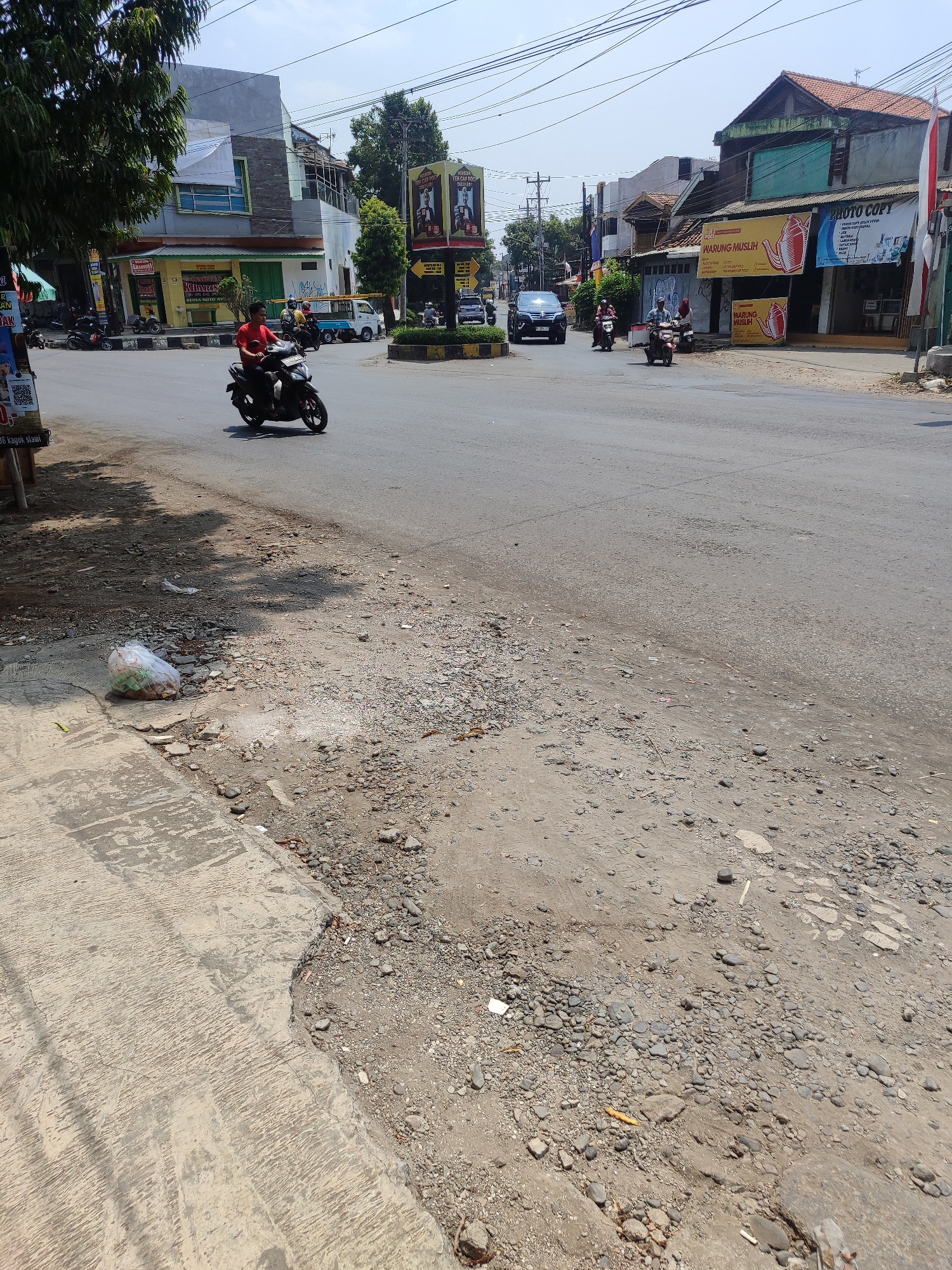 The image depicts a street scene in Kagok, Indonesia, on a bright, sunny day. The road surface in the foreground is uneven, with patches of gravel and dirt, suggesting it may be undergoing repair or is in a state of disrepair. A concrete sidewalk runs along the left side, with a large bag of trash lying on its edge.
In the midground, a man in a red shirt is riding a black motorcycle towards the right of the frame. Further down the road, several other motorcycles and a small white truck are visible, indicating moderate traffic. Buildings line the street on both sides. On the right, several shops are identifiable by banners, including one with "WARUNG MUSLIM" written on it. The buildings are a mix of styles, with some appearing older and others more modern. Overhead, electrical wires are strung across the sky.
In the background, more buildings and trees are visible, along with a roundabout in the distance, suggesting this is a crossroads or intersection. The overall impression is of a typical urban or semi-urban environment in Indonesia, with bustling activity and the characteristic elements of Indonesian street life.
