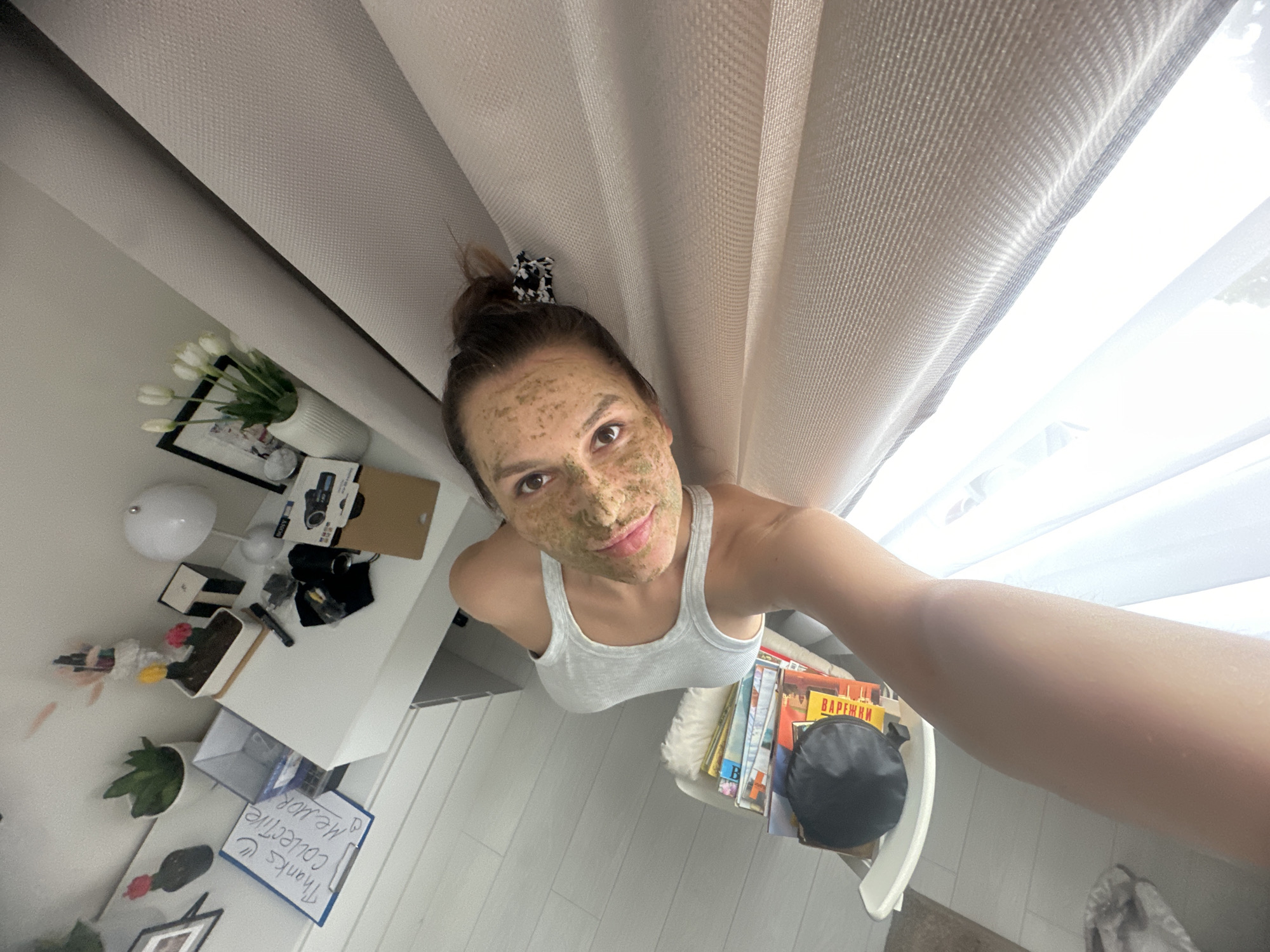 This high-angle selfie captures a young woman from directly above, creating an unusual perspective. The woman, with brown hair styled in a messy bun secured by a black and white checkered scrunchie, has a green face mask applied to her entire face. She is wearing a light grey ribbed tank top and looks directly at the camera with a neutral, calm expression, a faint smile on her lips. Her right arm is extended, holding the camera for the selfie.
The "ceiling" or upper part of the image features light-colored, textured curtains, with a sheer white curtain behind them allowing bright natural daylight to enter, indicating it is daytime.
On the left side, a white table or shelf is cluttered with various objects: a white spherical lamp, a black-framed picture, a white fluted vase containing artificial white tulips, small boxes (one black), camera equipment including a black camera body and lenses, and a small green potted plant. A clipboard with visible text, "Thanks U! Collective me, Mor," is also present.
On the right, another white surface holds a stack of books. The spine of one book prominently displays "ВАРЕЖКИ" (Varezhki - Mittens) in Cyrillic, a strong location cue supporting Moscow, Russia. A black, soft item, possibly a bag, rests on the books. The "floor" visible at the bottom of the image is light-colored, likely wood or laminate, with visible planks.
The activity depicted is a personal self-care or beauty routine, with the woman applying a face mask and documenting it. The scene conveys a casual, domestic setting.