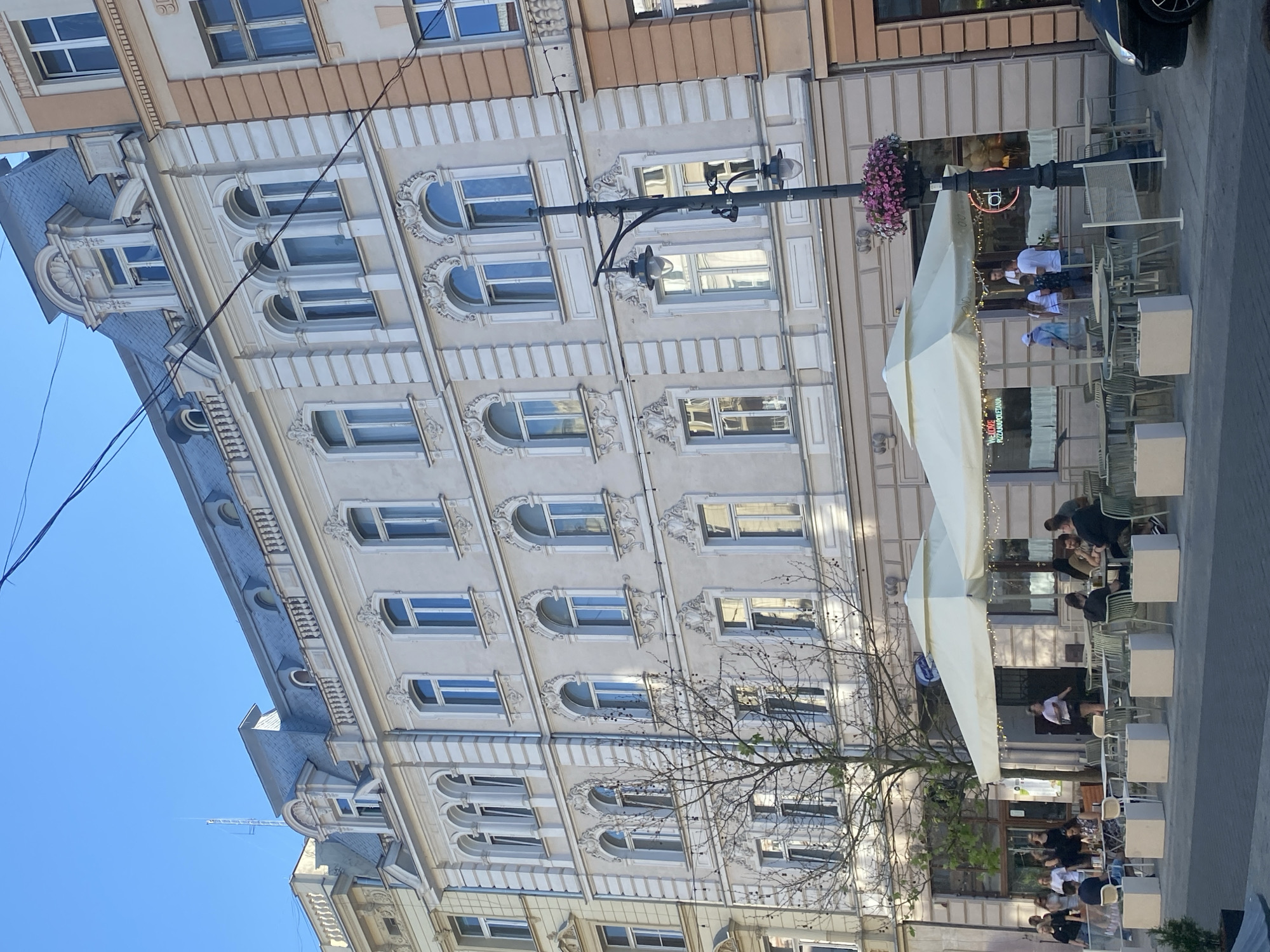 This media file captures an outdoor scene in front of a building with an ornate facade, likely a cafe or restaurant. Several people are seated at tables, some are standing and walking. The time of day appears to be late afternoon or early evening, as indicated by the soft lighting and shadows. The weather seems pleasant, conducive to outdoor dining.
The building's architecture is detailed, featuring a series of windows with arched tops and decorative trim. A street lamp with two lights stands prominently in the foreground, adorned with a cluster of pink flowers. A large, light-colored awning extends over some of the outdoor seating area.
Visible text includes "PIZZA NEAPOLITANA" on a sign. The presence of multiple people engaged in dining and socializing suggests a relaxed and leisurely atmosphere. The location context of Łódź, Poland, provides a geographical reference for the scene.