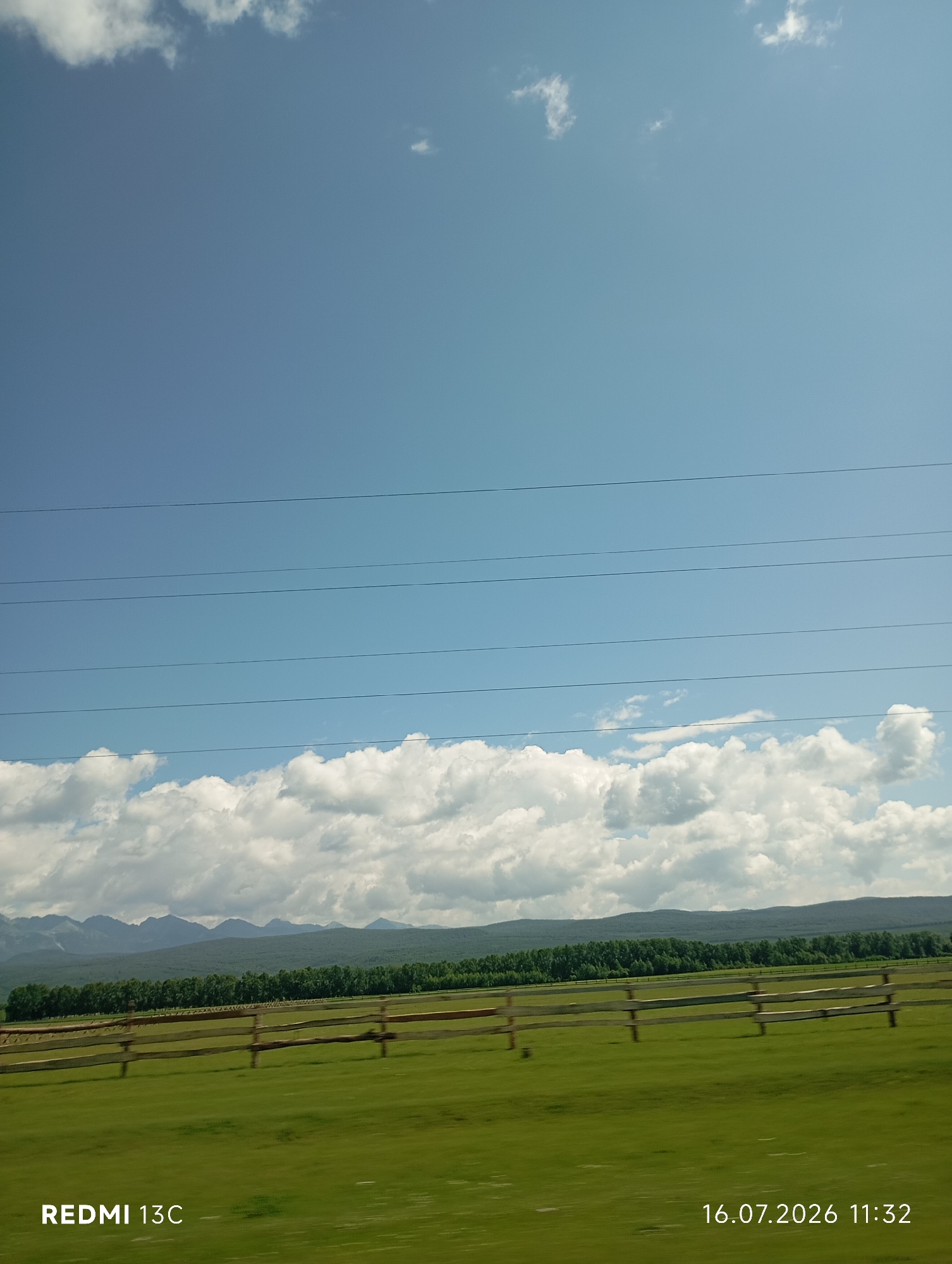 An exterior, medium-shot photograph captures a wide, grassy field bordered by a wooden fence, with a dense line of green trees behind it. In the distance, a range of muted, blue-gray mountains forms the horizon. Above the trees, a band of white, puffy clouds stretches across a bright blue sky. Several dark, horizontal power lines traverse the sky. The bottom left corner of the image displays the text "REDMI 13C," and the bottom right corner shows the date and time, "16.07.2026 11:32." The scene is set in Galbai, Russia.
