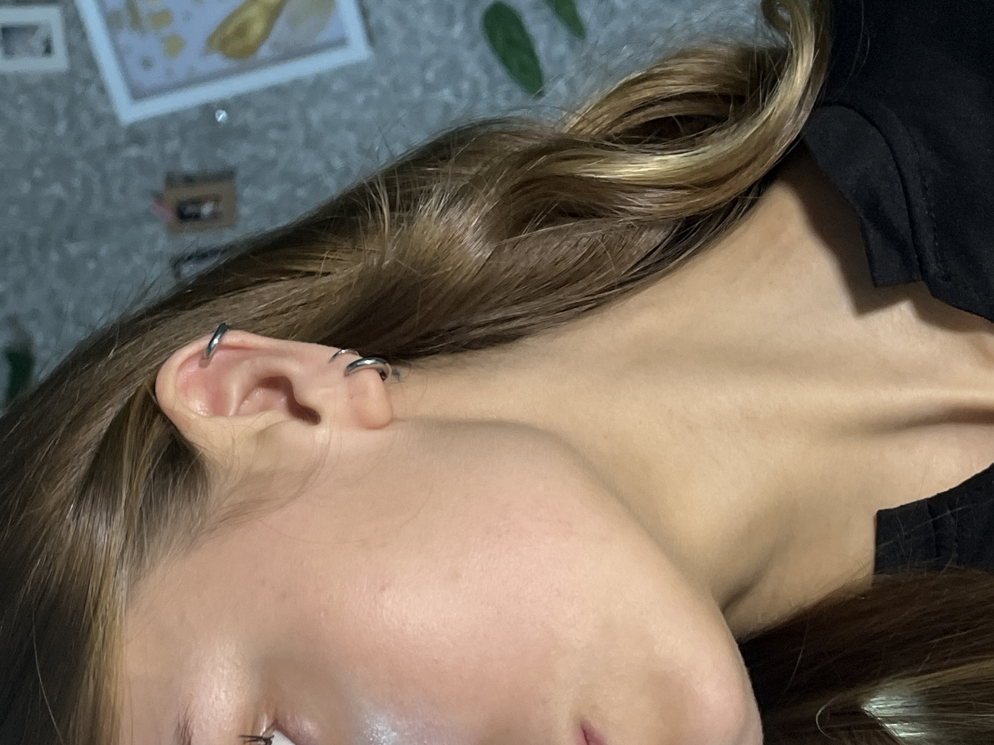 A close-up, angled view of a person's head and neck. The person has light skin, and their hair is brown with blonde highlights. They have at least two silver hoop earrings in their left ear. The background includes part of a patterned surface and an out-of-focus picture on the wall. The person is wearing a black garment. There is no indication of the location being Фоки, Russia.