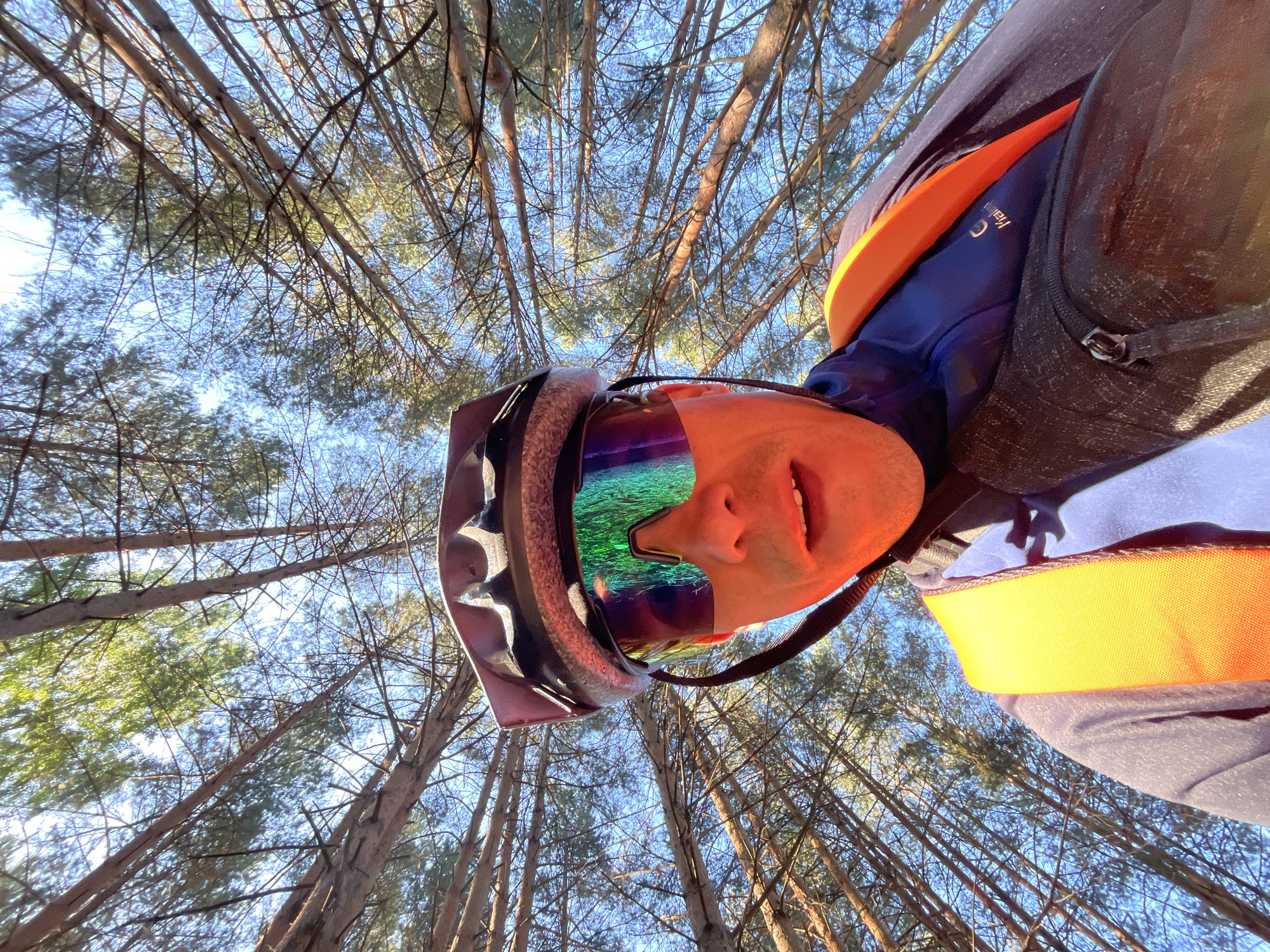 A low-angle, wide shot shows a person wearing a dark helmet and iridescent cycling glasses looking up at the camera. They are outdoors, surrounded by tall trees with a bright blue sky visible through the branches. The person is wearing a dark blue jacket with an orange strap visible across their chest and a black fanny pack around their waist. The image captures a moment of a cyclist in a wooded area, likely in the city of Chaikovsky, Russia, given the context.