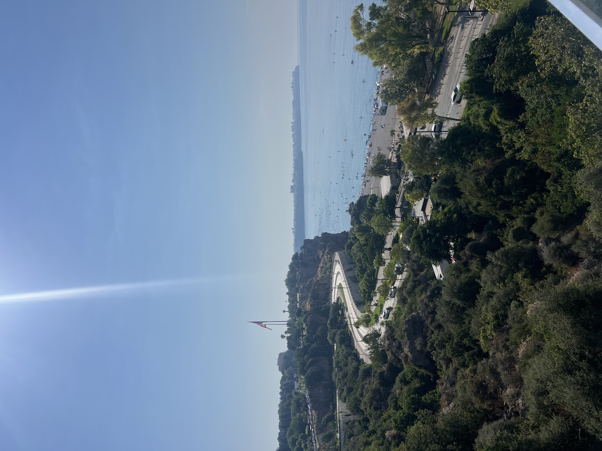 This is a high-angle, eye-level, wide shot photograph taken on a sunny day. The image depicts a scenic coastal road curving along a cliff face, with lush greenery on the left and a bustling beach and the Mediterranean Sea on the right.
The foreground is dominated by dense, dark green foliage, with some lighter green and dry shrubbery interspersed. A winding road with white lane dividers and streetlights is visible, carved into the side of the cliff. Several cars are driving on the road, including a white sedan in the mid-ground. Further up the cliff, a large Turkish flag flies from a tall flagpole, with another smaller flag visible on a nearby pole. Buildings and more trees are scattered along the top of the cliff.
To the right, a sandy beach is packed with numerous people sunbathing, swimming, and engaging in various beach activities. Umbrellas and beach chairs are visible. The sea is a calm, light blue, with several small boats and individuals on paddleboards or kayaks. A distant city skyline stretches across the horizon. The sky is a clear, bright blue with a strong sun flare emanating from the upper left corner. The overall impression is a vibrant, lively scene at a popular tourist destination. The location appears to be Antalya, Turkey, likely overlooking Konyaaltı Beach or a similar coastal area.