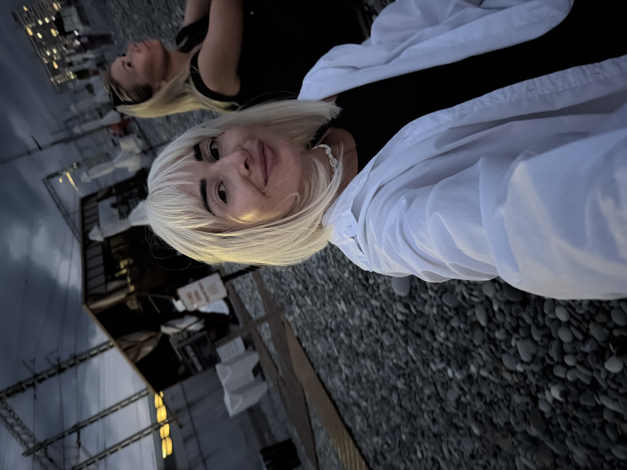 The image depicts two women on a pebble beach in Sochi, Russia, near dusk. The woman in the foreground, with blonde bob-cut hair and bangs, is looking towards the camera and smiling. She is wearing a white unbuttoned jacket over a black top and a shell necklace. The second woman, with blonde hair pulled back and sunglasses on her head, is visible in the background to the right, looking upwards. She is wearing a black top. The background shows a dark, overcast sky, structures resembling a kiosk or small building, and what appear to be power lines or train tracks. The ground is covered with dark gray pebbles. The overall lighting suggests it is either early morning or late evening.