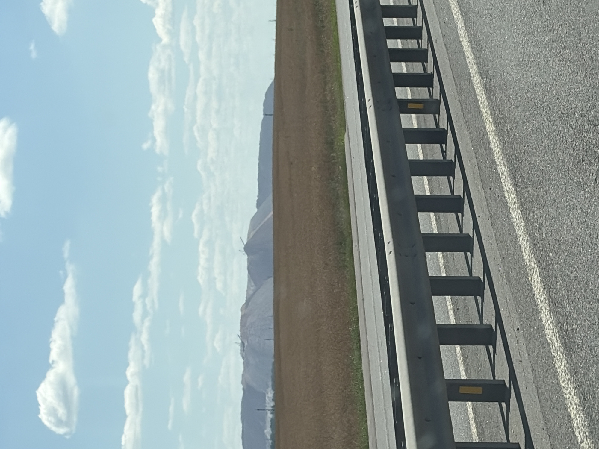 The image depicts a wide, flat landscape under a bright blue sky with scattered white clouds. In the foreground, a paved road with white lane markings is visible, separated by a metallic guardrail. The guardrail consists of vertical posts supporting a horizontal beam, with yellow rectangular reflectors attached to some of the posts. Beyond the road and guardrail lies a field of dry, golden-brown grass or crops. In the distance, a range of large, pale mounds dominates the horizon. These mounds appear to be artificial, possibly waste piles from mining or industrial activity, with several cranes or heavy machinery visible on their slopes. The overall impression is one of an industrial or agricultural area with a wide-open sky. The location is identified as being within the Belitski Sel'ski Sovet, Belarus.