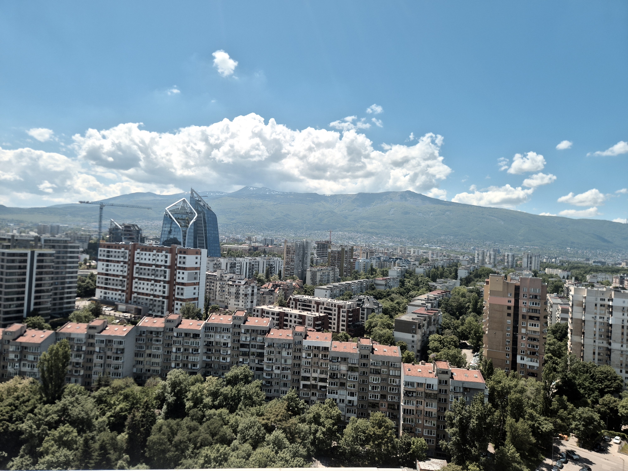 The image captures an elevated view of Sofia, Bulgaria, on a sunny day with a clear blue sky dotted with fluffy white clouds. In the foreground, there's a dense urban area consisting of residential buildings with tiled roofs partially obscured by lush green trees. Mid-ground features a modern skyscraper with a distinctive, crystalline design, and other high-rise buildings in various architectural styles. In the background, a mountain range, Vitosha, stretches across the horizon, its peak partially covered with patches of snow. A construction crane is visible near the modern skyscraper, indicating ongoing development in the area. The city appears densely populated with a mix of old and new architecture.