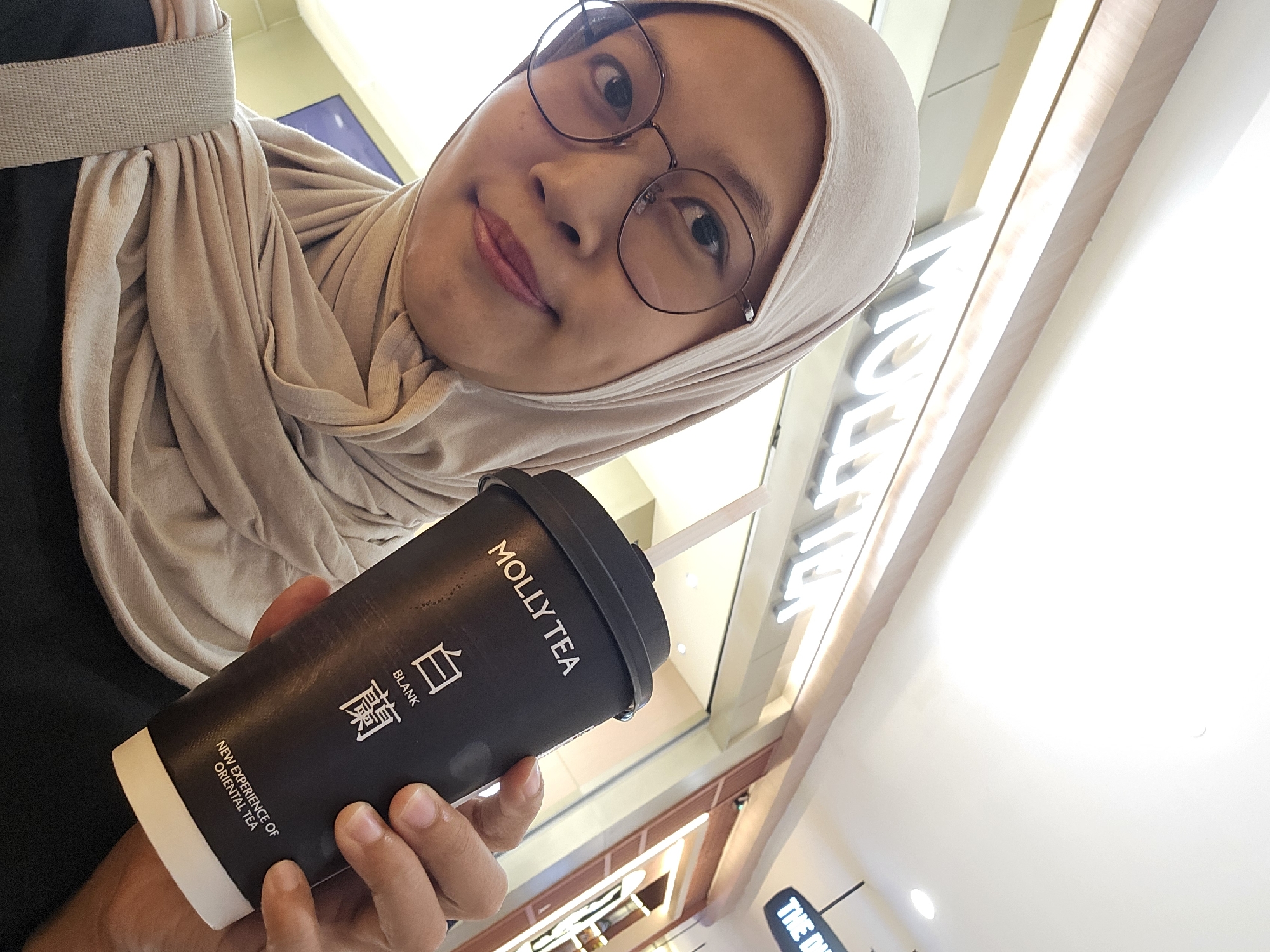 A young woman wearing a light beige hijab and glasses smiles at the camera while holding a black "MOLLY TEA" cup. The cup has white text and Chinese characters on it, along with the phrase "NEW EXPERIENCE OF ORIENTAL TEA". In the background, a store with illuminated signage that reads "MOLLY TEA" is visible, suggesting the location is likely in a mall or commercial area in Condongcatur, Indonesia. The overall lighting suggests an indoor setting.