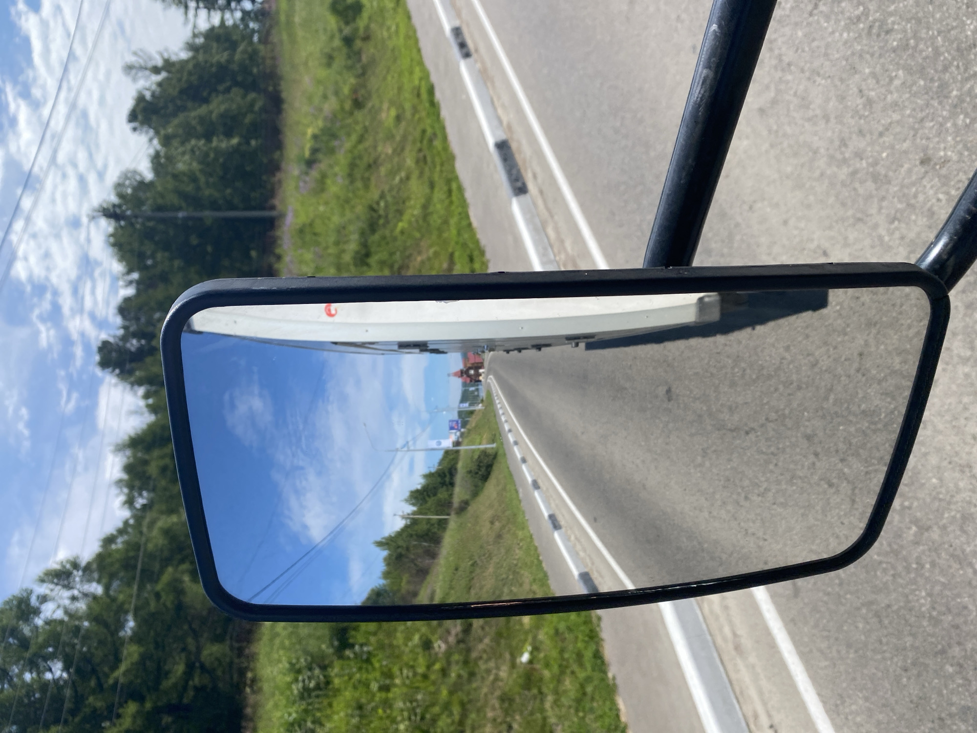 The image shows a reflection in a side-view mirror of a vehicle. The reflection shows a road with a white and grey striped curb, a grassy slope with trees on the left, and a clear blue sky with clouds. In the distance, a red-roofed building, possibly a church, is visible. The road appears to be in a semi-rural area. The scene is captured from the perspective of someone inside a vehicle, looking at the reflection in the mirror. The setting is consistent with the location in Listvyanka, Russia, a village on the shores of Lake Baikal, known for its scenic beauty and natural landscapes. The image depicts a typical view one might encounter while traveling on a road in such an area.