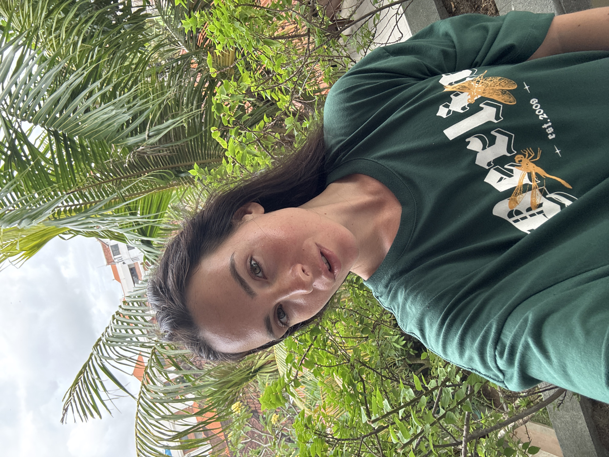 A dark-haired woman is taking a selfie outdoors. She is wearing a dark green t-shirt with white lettering and two dragonfly graphics. Behind her, palm fronds and lush green foliage are visible, with buildings in the background. The sky is overcast. The scene appears to be located in North Kuta, Indonesia.