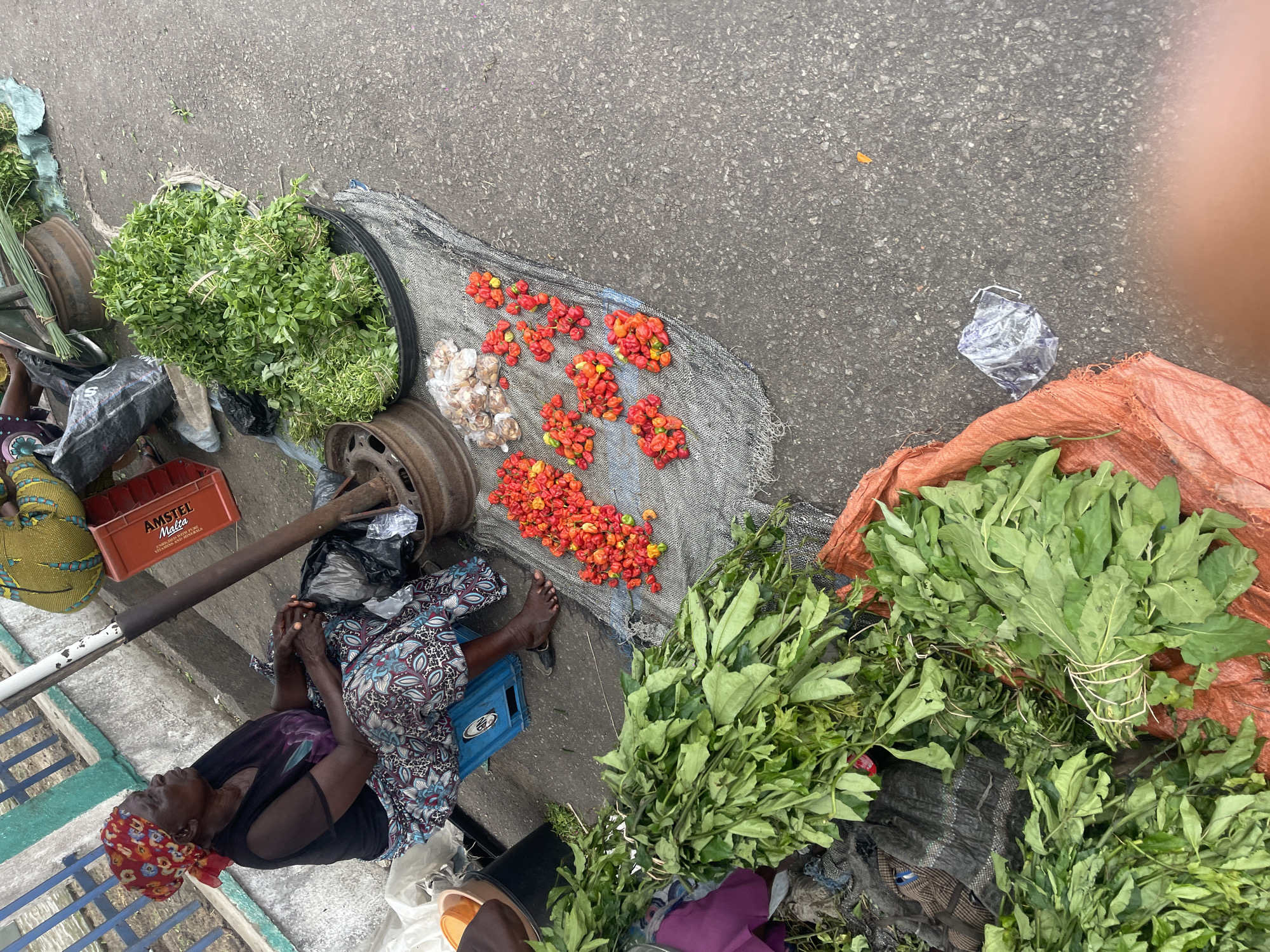 An overhead shot captures a vibrant outdoor market scene, likely in a West African setting. An elderly woman, possibly a vendor, is seated on a blue plastic crate, resting with her hands clasped. She wears a colorful red headscarf with floral patterns, a dark top, and a patterned skirt, her bare feet visible.
Around her are various fresh produce items displayed on the asphalt ground and on tarps. To her right, large bundles of dark green leafy vegetables are arranged on an orange tarp and a black mesh cloth. Directly in front of her, on the black mesh, are multiple piles of small, bright red and orange peppers, alongside small clusters of white produce, possibly garlic or onions. Further to the left, in a black woven basket, are smaller bundles of light green, round-leaved herbs or vegetables.
Other objects visible include a rusty metal wheel rim, a dark metal pole, and various plastic bags. An empty red plastic crate, prominently displaying "AMSTEL MALTA" and partially "VITAMINS AND MINERALS," lies near her. On the far left, bundles of what appear to be straw brooms or grass are stacked. The scene is illuminated by bright, natural daylight, indicating it is daytime. The overall impression is that of an active, informal market stall.
