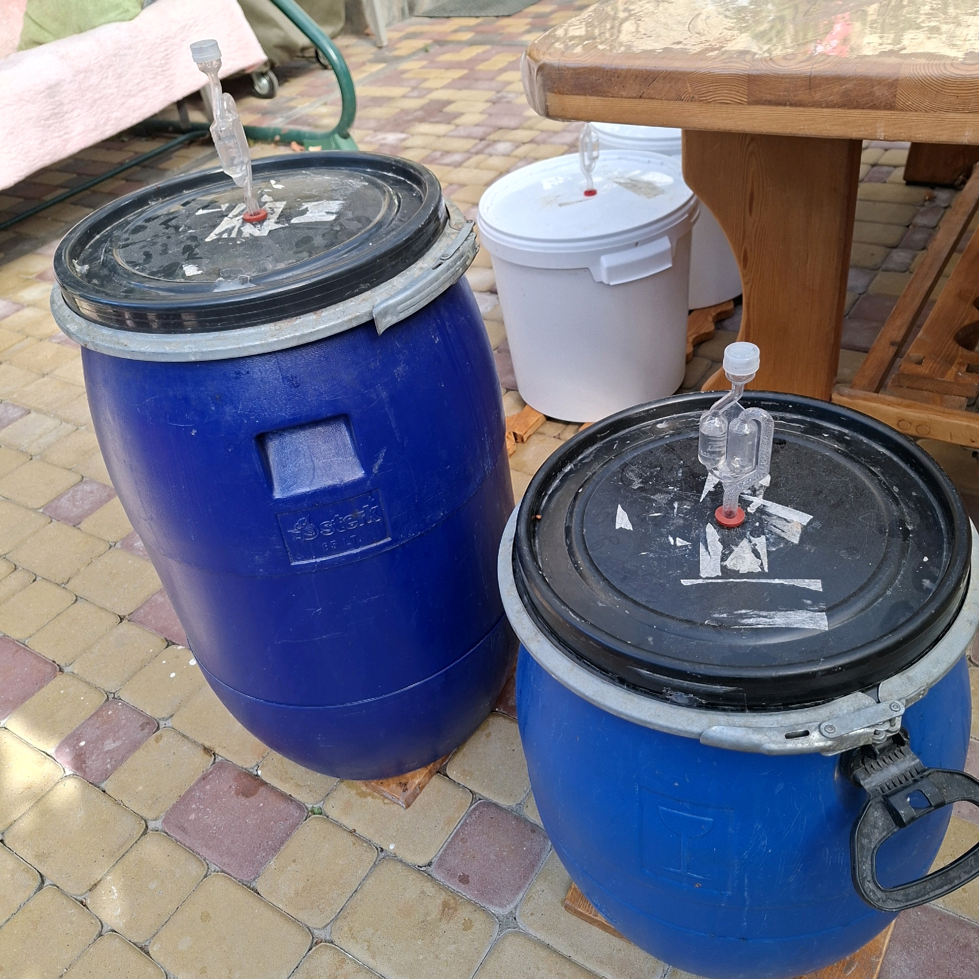Two blue plastic barrels with black lids are positioned outdoors on a paved patio. Each barrel has a clear plastic fermentation airlock installed in its lid. To the right of the barrels is a wooden table with a light-colored top. In the background, a white plastic bucket with a white lid is visible, also with a fermentation airlock installed. A pink blanket or tarp is partially visible in the upper left corner, draped over what appears to be a piece of furniture. The scene is set on a sunny day, indicated by the lighting and shadows.