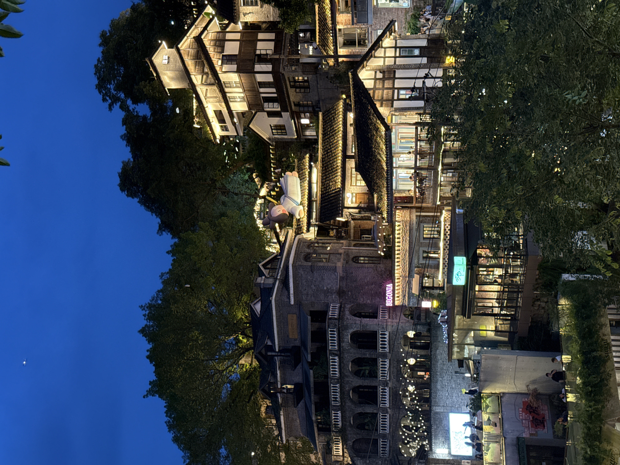 The image depicts a night scene in Nan'an District, Chongqing, China. Several multi-story buildings of mixed architectural styles are visible. These include traditional Chinese structures with dark tiled roofs and exposed timber frames, and more modern buildings with arched windows and facades of brick and stone. The buildings are illuminated with warm, artificial light. Trees line the perimeter of the scene, bordering the view from the blue night sky. A restaurant sign reading "BEGONIA" is prominent in neon pink. A street is seen, occupied by groups of people. String lights are strung between structures. There are trees and greenery in abundance. In the mid-ground, a large, cartoon character is suspended from the roof.