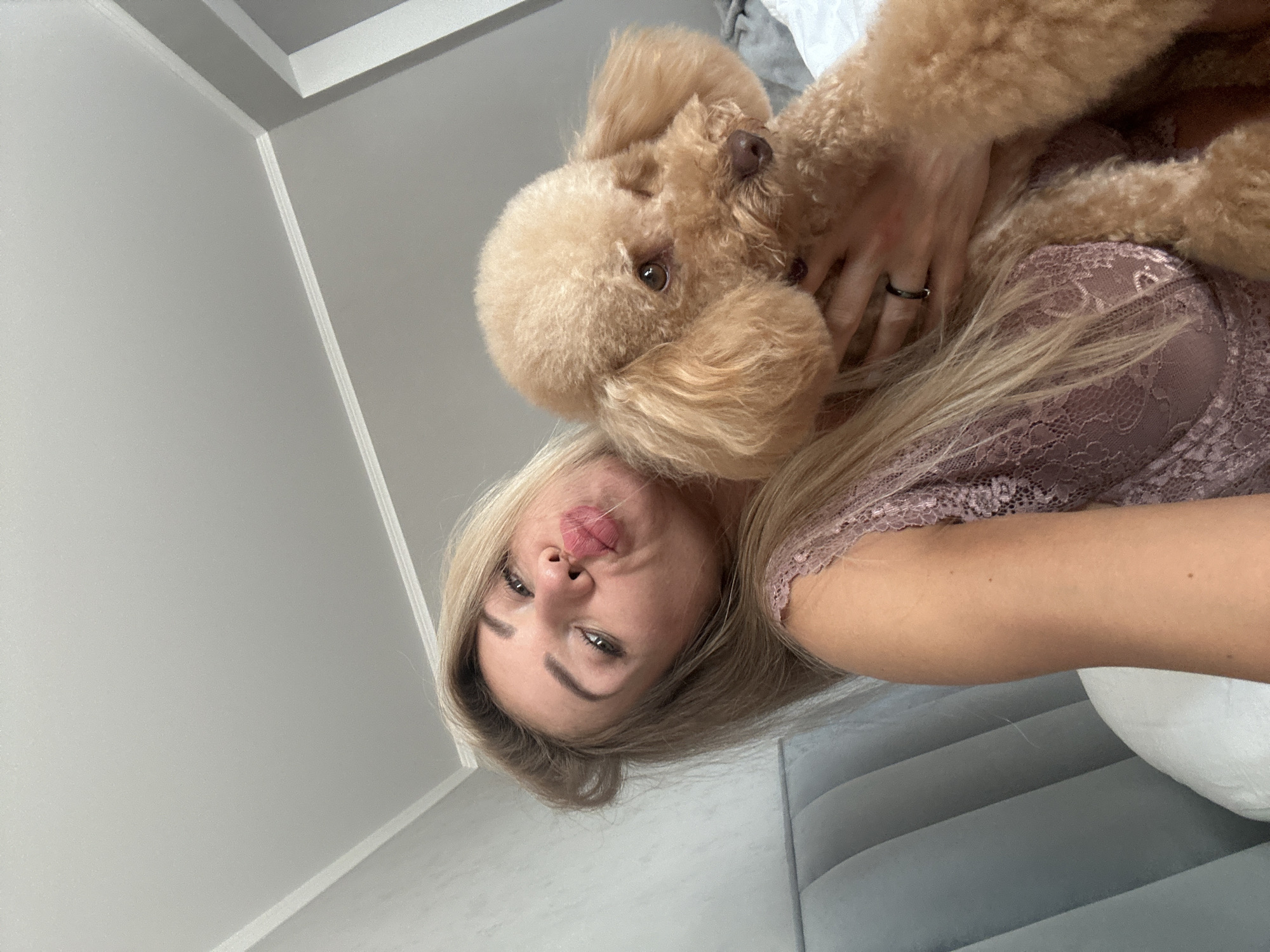 A blonde woman wearing a pink lace top is taking a selfie in bed, holding a light brown poodle. The woman is puckering her lips for the camera. She has long, wavy blonde hair. The poodle is being held close to her face, with its head turned to the side. The background shows a grey upholstered headboard and a white pillow. The overall impression is of a casual, intimate moment. The image does not contain any specific geographical markers that would indicate its location in Armavir, Russia.