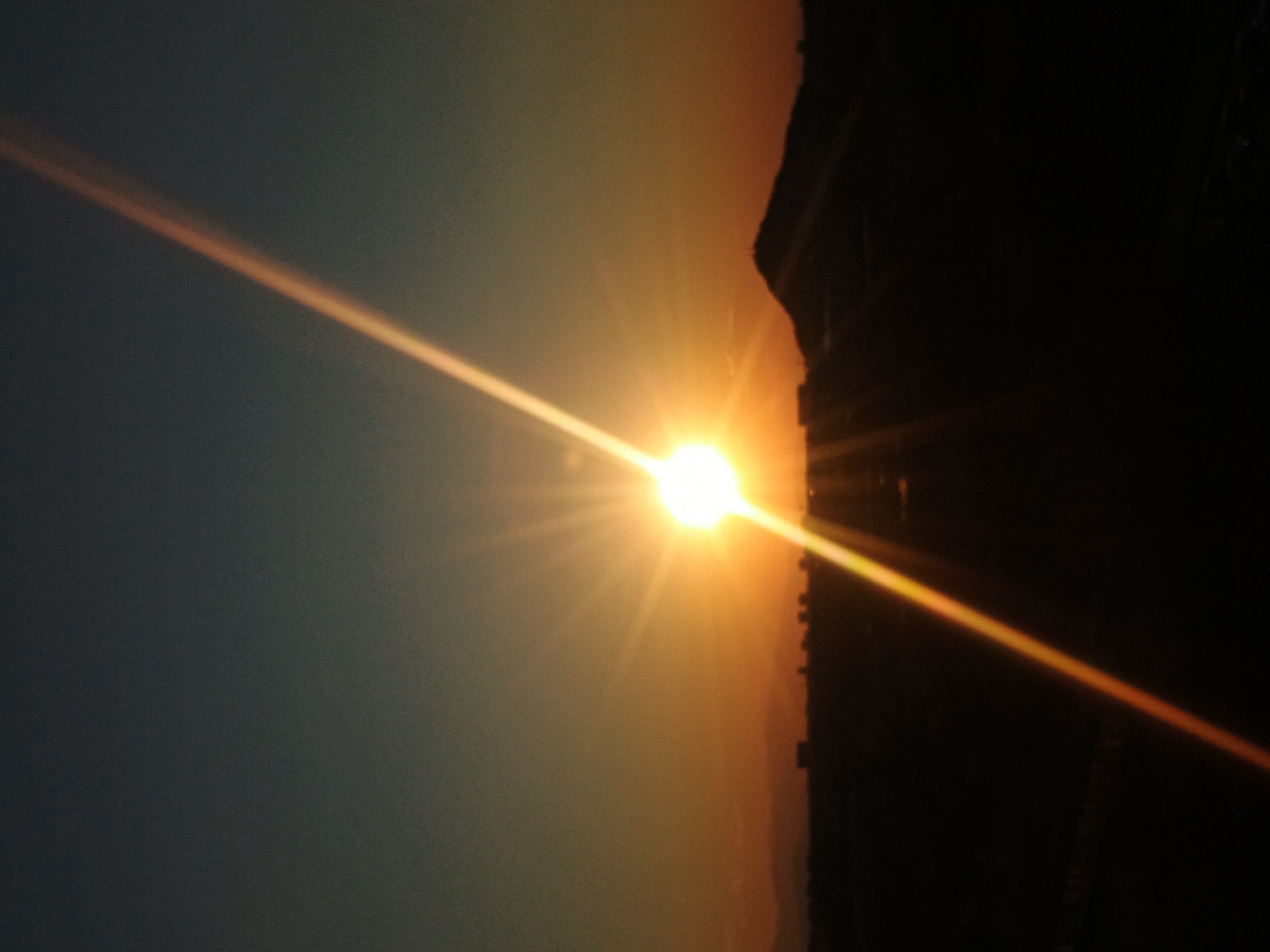 The image shows a bright sun low on the horizon, casting a strong glare and rays of light across the scene. To the right of the sun is a dark, silhouetted building or structure, possibly part of a cityscape. The left side of the image consists of a dark blue-gray sky, fading into a gradient of orange near the horizon where the sun is located. Given the context of Plovdiv, Bulgaria, the scene could be a sunset over the city, with the silhouette possibly representing buildings in the central area.