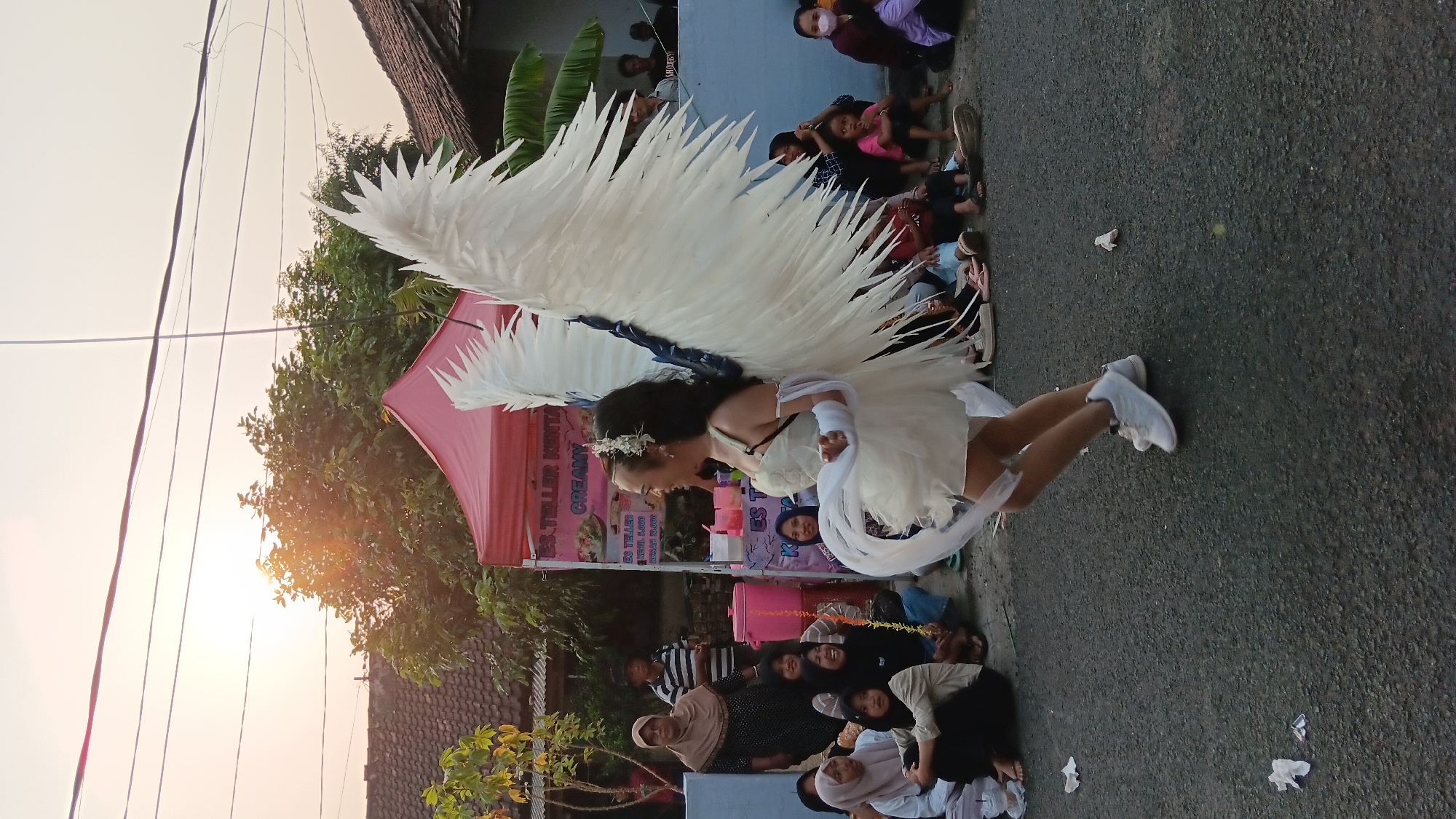 In Cerme, Indonesia, an individual adorned with large white angel wings and a light-colored, short dress is the central subject of this image. They are captured mid-stride on an asphalt road, their body turned to the left, face looking towards the left. A white sash is draped from their right hand. The event appears to be outdoors, with onlookers seated on the ground and along a low wall to the right and left of the road. Behind the seated figures, a pink tent with text advertising "ES TELER KENTANG CREAMY" is visible, suggesting a local market or fair. The background features residential buildings with tiled roofs and trees, under a bright, hazy sky with the sun casting a warm glow. The overall scene depicts a community gathering or street performance.