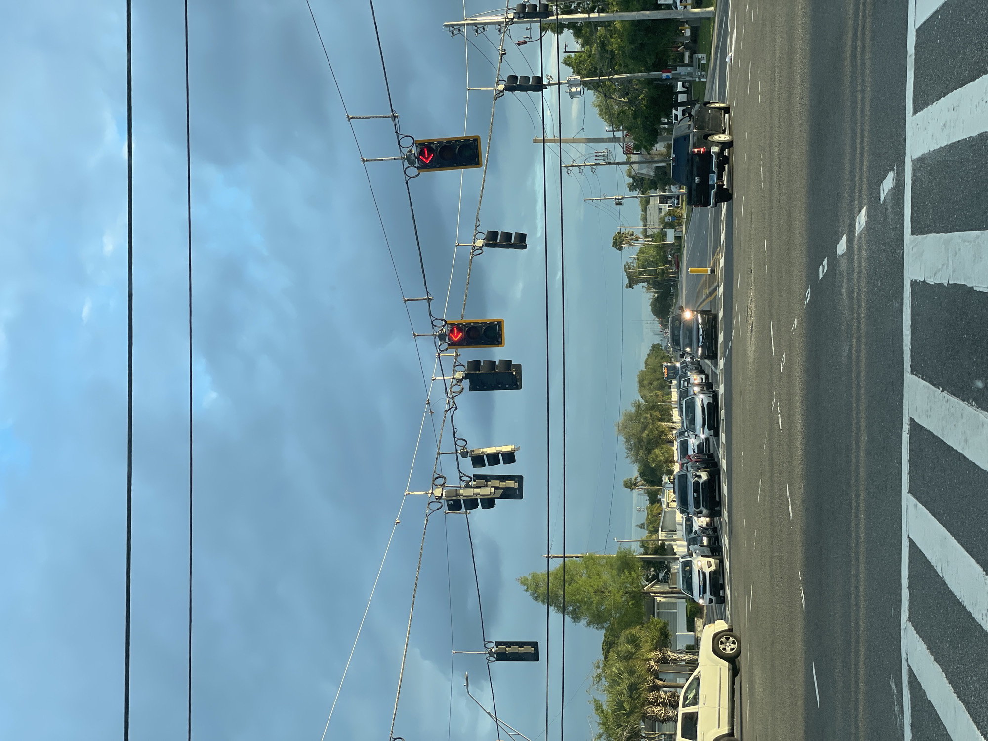The image depicts a street scene in Largo, Florida, United States of America. The view is likely from inside a vehicle. The street is lined with multiple vehicles stopped in traffic. Overhead, several traffic lights are visible, all displaying red arrows. The sky is overcast with dark clouds. Vegetation and buildings line the roadside. Crosswalk markings are visible on the road. Power lines are strung across the sky.