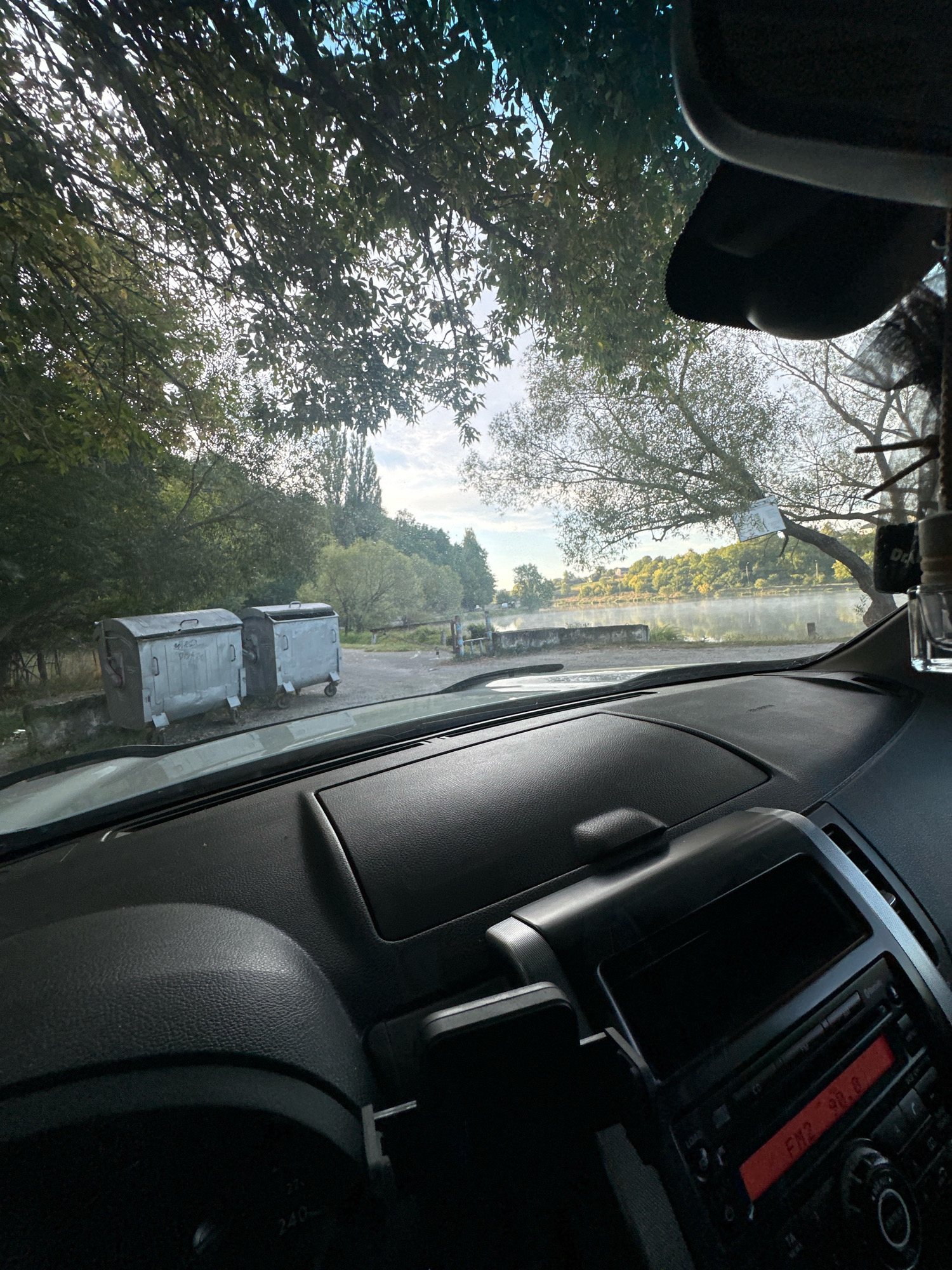 The image is taken from inside a car, looking out through the windshield. The dashboard of the car is visible in the foreground, with the steering wheel and radio. Outside the car, there are two large, grey garbage bins on wheels, situated on a gravelly surface. Beyond the bins, a body of water, likely a lake or river, is visible, with trees and vegetation lining its banks. The sky is overcast with some patches of blue. The overall scene appears to be in a rural or park-like setting, possibly near a recreational area. The text on the radio display reads "FM2 98.8". The image is described as being located in the city of Makiv, Ukraine.