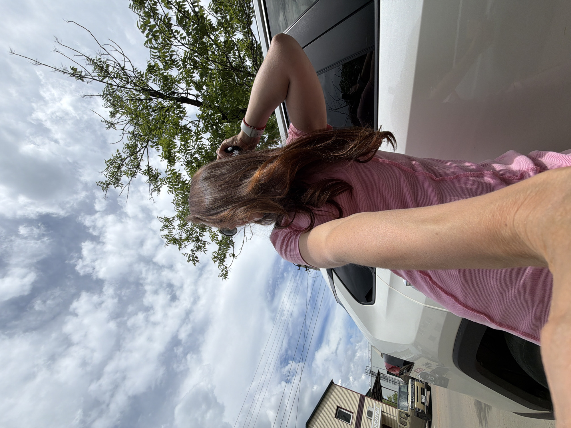 A woman with long, dark brown hair is captured in a selfie-style shot, standing outdoors next to a white SUV. She is wearing a pink ribbed t-shirt and has a white fitness tracker on her left wrist. Her right arm is bent at the elbow, and she appears to be adjusting something near her ear with her hand. The background shows a cloudy sky with patches of blue, a green tree with visible branches, overhead power lines, and a building with light-colored siding and a dark roof on the left side of the frame. A small, light-colored car is parked in front of the building. The reflection in the SUV's window shows a faint image of the woman and potentially another person. The scene appears to be in an urban or suburban area, likely in Birobidzhan, Russia, given the context.