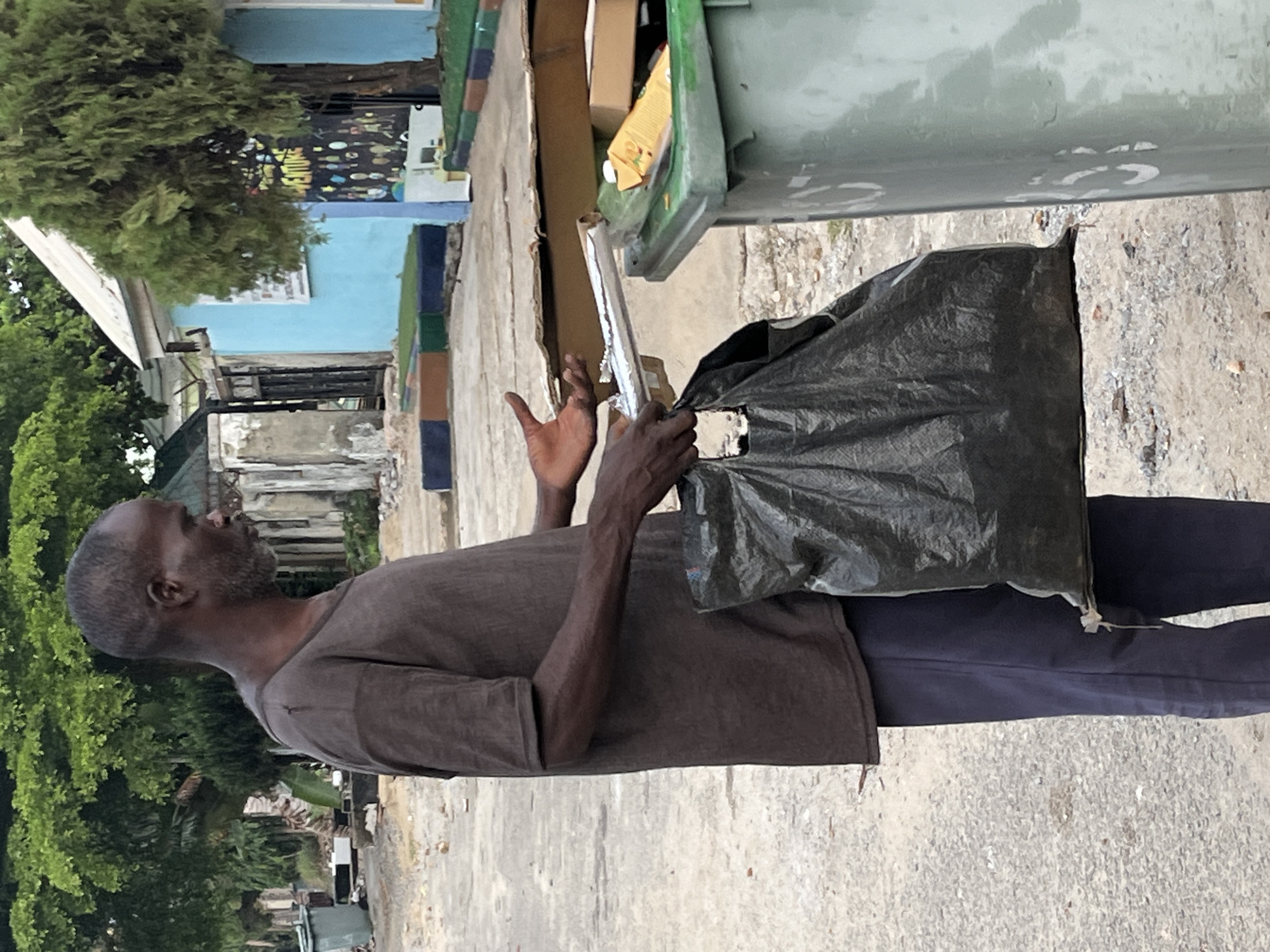 A man in a brown t-shirt and dark pants is seen putting a long, silver object into a trash bag that he is holding open. The trash bag is next to a green trash can, and there are boxes inside. He is outside in a neighborhood in Abuja, Nigeria. The man has gray hair and a beard. The background includes trees, a blue building, and a street.