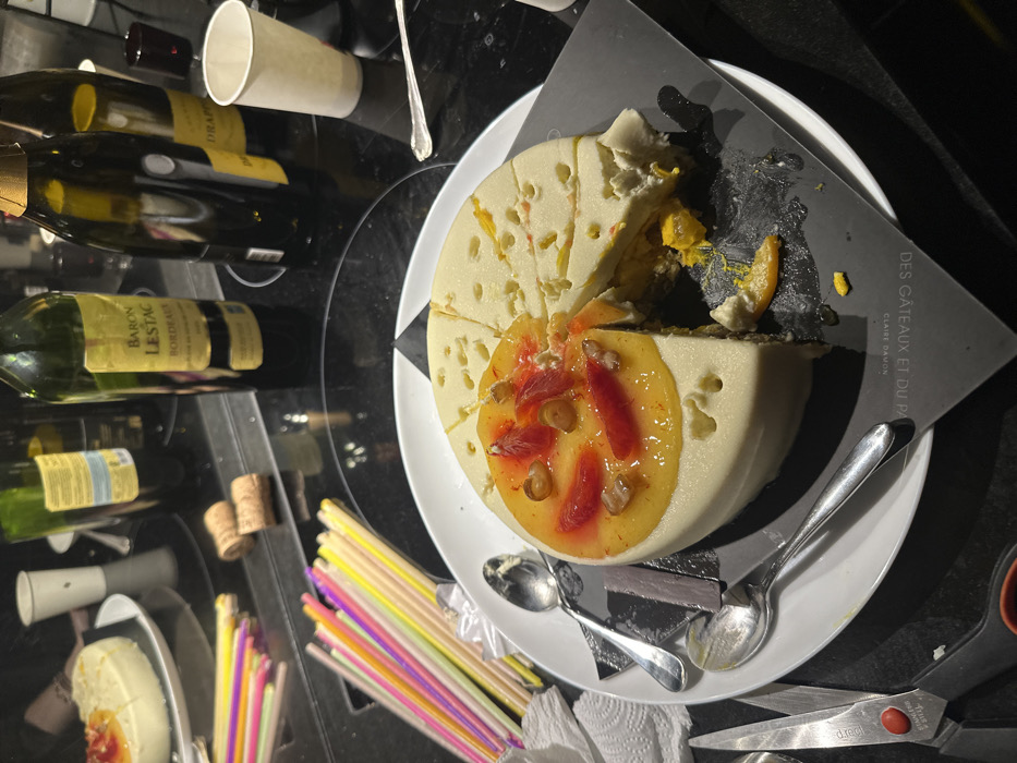 The scene depicts an indoor table setting with food and beverages, indicating consumption from "Des Gâteaux et du Pain" by Claire Damon, identifying the location as Paris, France.
A round, light-colored cake, featuring a perforated outer layer and topped with a yellow-orange glaze, sliced red fruit (likely blood orange), and small brown nut pieces, is presented on a white plate over a black paper bearing the patisserie's name. A single slice of the cake has been removed, and adjacent portions show signs of disturbance, revealing a yellow-orange interior filling. Three silver spoons are positioned around the cake: two on the plate, one near the left edge.
To the immediate left of the cake, a cluster of multi-colored plastic straws (yellow, green, pink, purple) rests beside a crumpled white paper napkin. A pair of silver-bladed scissors with red and black handles is visible in the bottom-left corner of the frame. Behind the straws, two wine corks are present.
The background displays multiple bottles on a dark, reflective surface. Prominently featured is a green wine bottle labeled "Baron Lestac Bordeaux." To its right is a dark champagne bottle with a gold foil top, and another dark bottle is further to the right. An additional green wine bottle is partially visible behind the Bordeaux bottle. A tall white paper cup is situated on the right side of the frame, with another white paper cup in the far left background. A second, smaller round dessert with a fruit topping is partially visible on a plate in the distant left background. The overall lighting is dim.