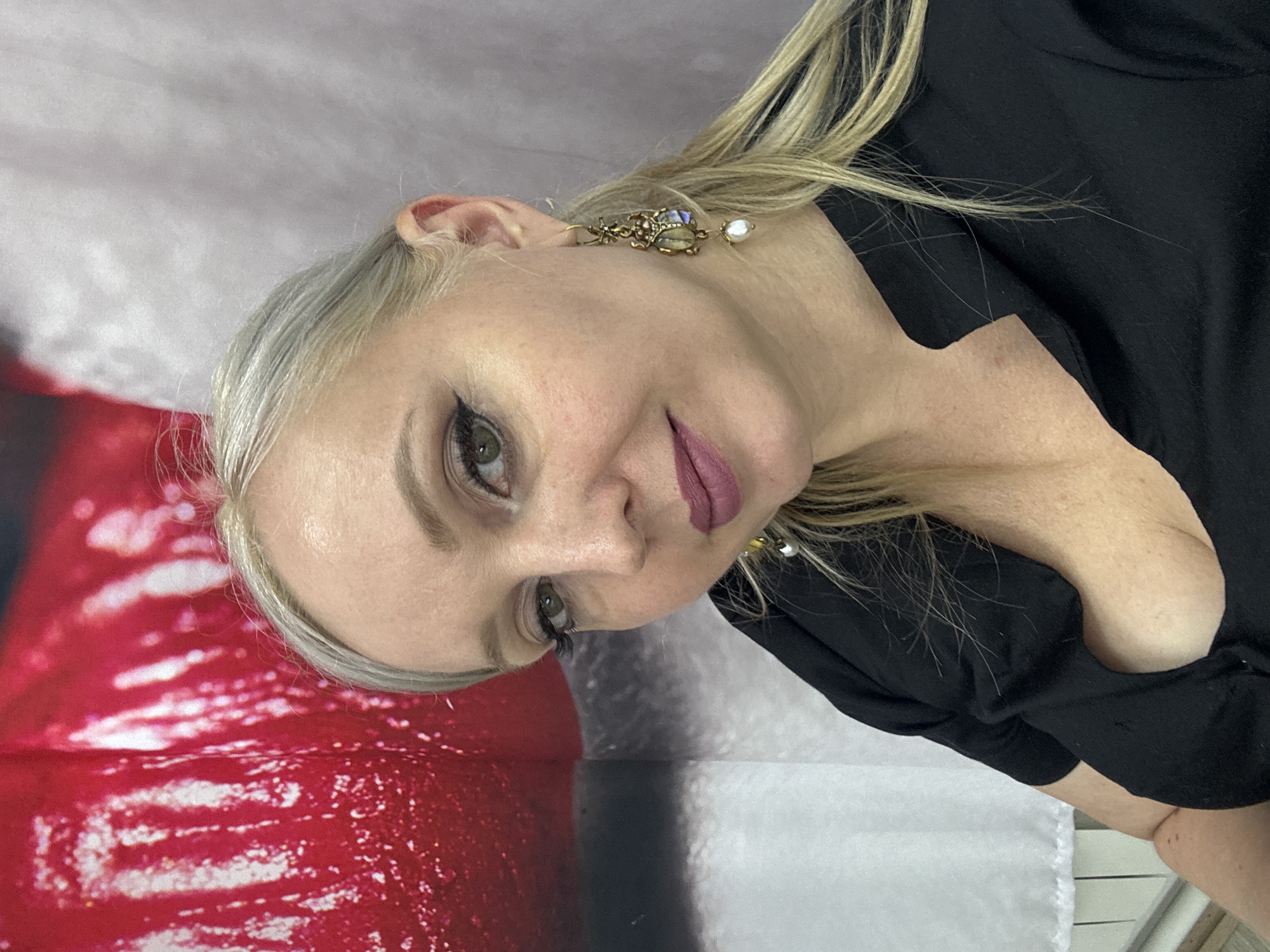 A blonde woman with green eyes and dark eye makeup is shown in a close-up portrait. She is wearing a black V-neck shirt and ornate dangle earrings with pearls and colorful gemstones. Her lips are painted a muted purple. The background consists of a blurred red and white abstract pattern on the left, and a textured greyish-white fabric on the right. The image appears to be a selfie taken indoors, possibly in a studio or residential setting in Komsomolsk-on-Amur, Russia, indicated by the provided metadata.