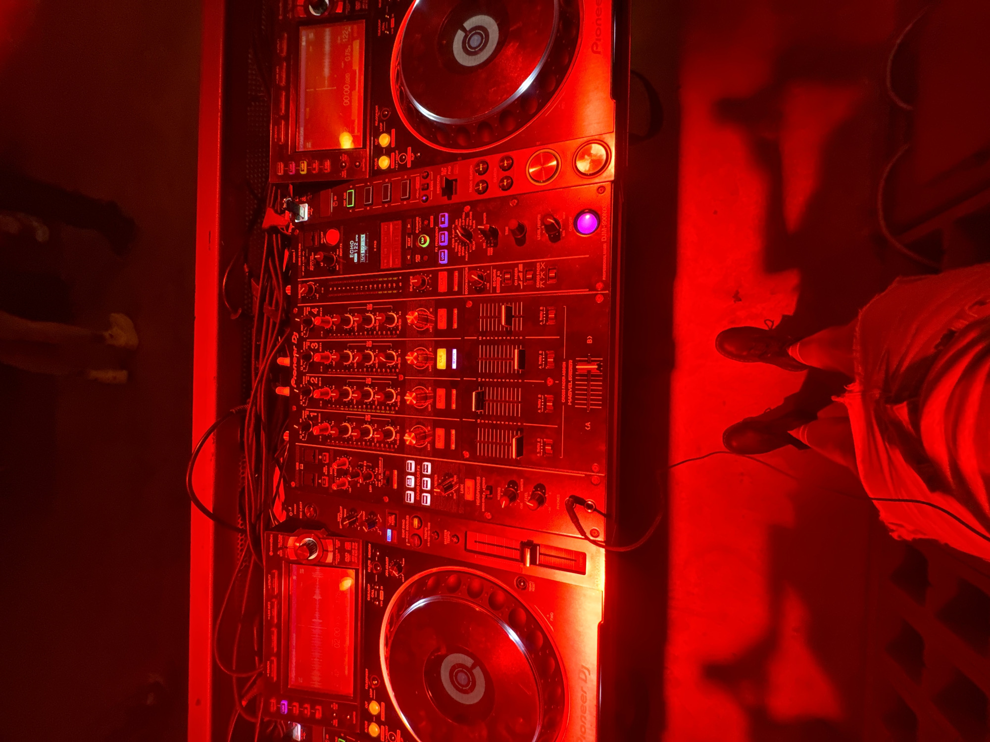 A DJ's equipment, consisting of two Pioneer CDJ decks and a DJM-900NXS mixer, is illuminated in red light. Cables connect the equipment. Partial views of people are visible to the left and right of the equipment. The location is not discernible from the image.
